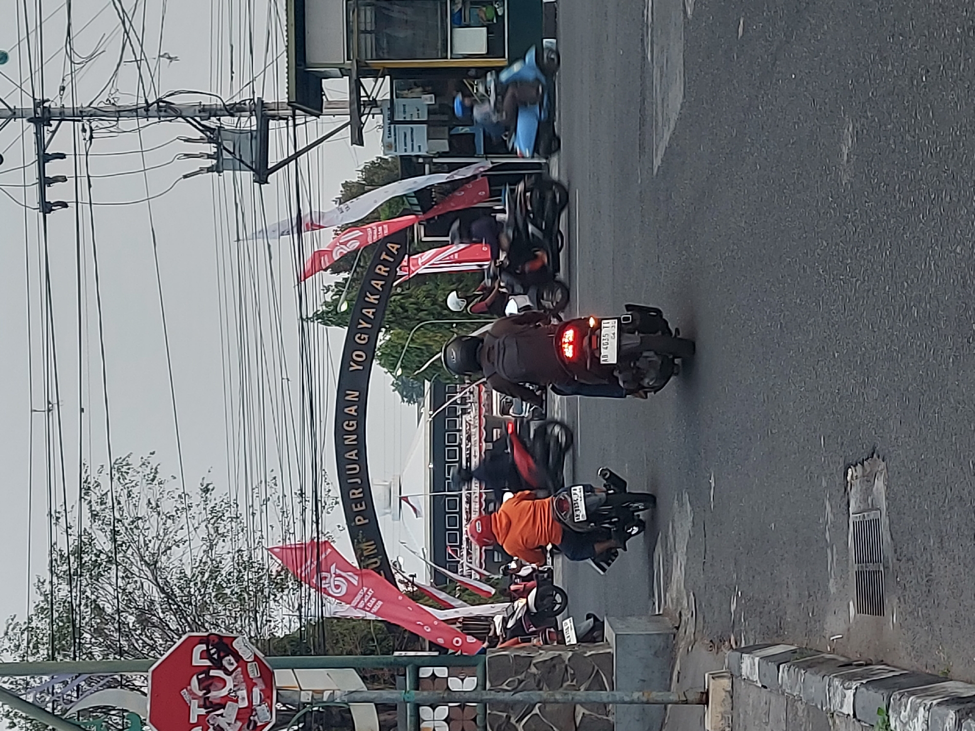 The image depicts a street scene in Yogyakarta, Indonesia, likely near the Museum Perjuangan Yogyakarta. In the foreground, several motorcycles are navigating the road. One motorcycle in the center, with a rider wearing a red helmet and an orange jacket, is moving away from the viewer. To its left, another motorcycle with a rider in an orange jacket and red helmet is also moving away. To the right, a blue scooter with a rider wearing a blue helmet is moving in the same direction. In the background, an archway with the text "MUSEUM PERJUANGAN YOGYAKARTA" is visible. Red and white flags are displayed, some bearing the text "95". A building with a white dome and dark windows is behind the archway. The environment appears to be an urban setting with trees, power lines, and traffic infrastructure. The sky is overcast.
