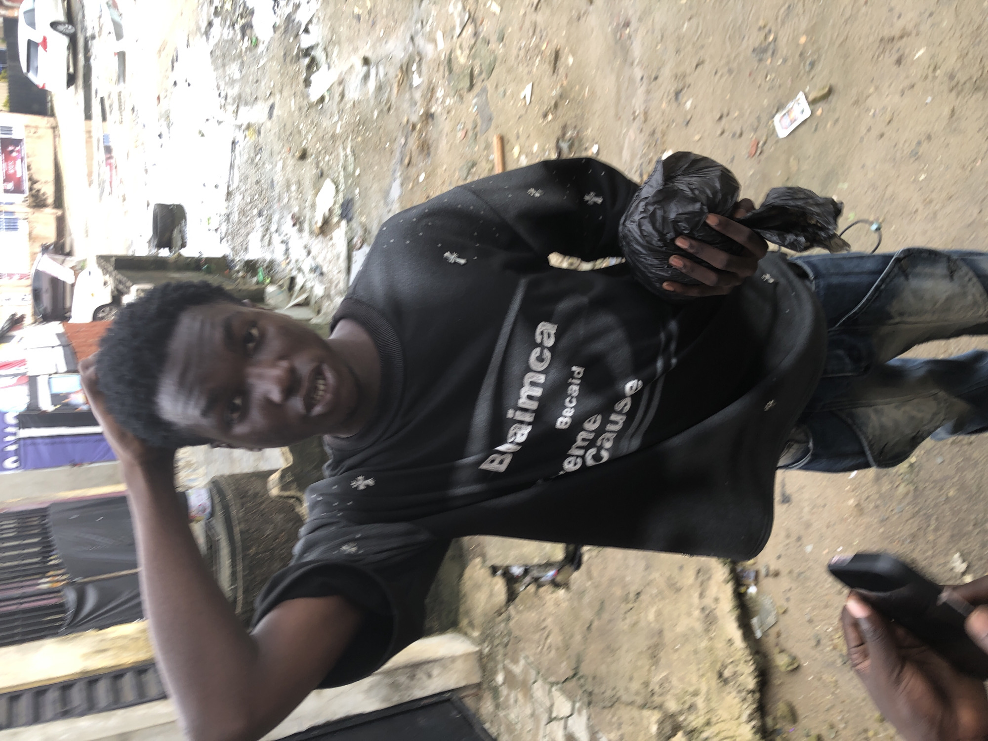 The media file shows a young Black male holding a black plastic bag in his right hand. He is wearing a black t-shirt with white text and small white cross designs. The text on his shirt appears to read "Blaimca" and "Becaideme Cause". He is also wearing blue jeans. His left hand is on the back of his head, and he is looking directly at the camera with his mouth open, showing his teeth. He appears to be smiling or speaking.
The background is a cluttered outdoor scene with dirt, debris, and some structures that suggest an urban or semi-urban environment. There are some posters or signs visible on buildings in the background. The ground is covered in dirt and what looks like scattered trash. The lighting suggests it is daytime. The overall impression is of a casual, informal setting. The individual's expression is lively and engaging. The context provided states the location is Ado, Nigeria.