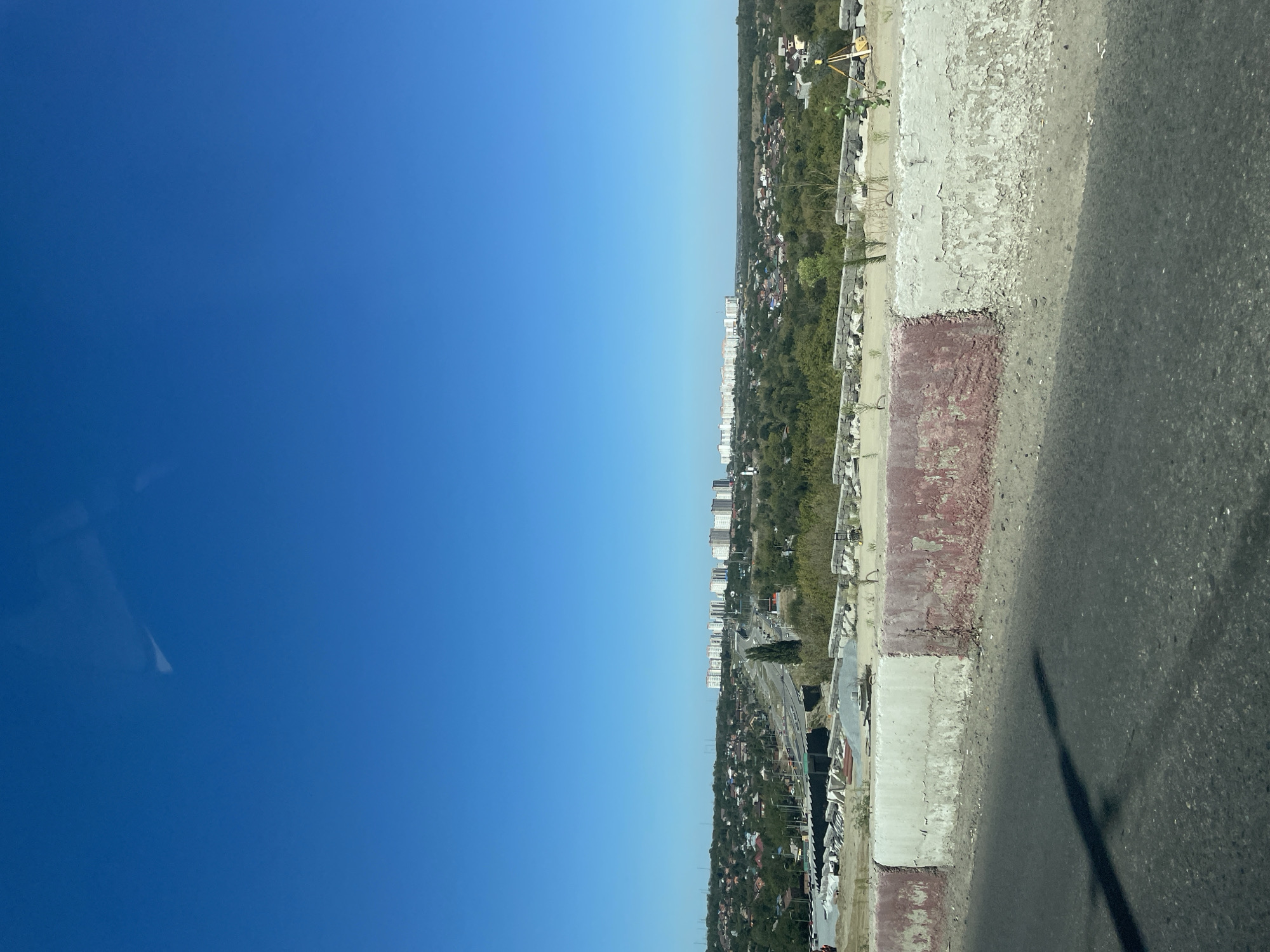 This is an eye-level, wide shot taken from a vehicle, looking out over a cityscape under a clear blue sky. In the foreground, a dark asphalt road is visible, with its surface showing a slight texture. To the right of the road is a low, weathered concrete barrier, painted with alternating sections of white and faded red. The white sections are peeling, revealing the concrete beneath. A shadow, possibly cast by the vehicle or a passenger, stretches across the road from the left side of the frame.
Beyond the barrier, the middle ground reveals a semi-urban landscape. There is evidence of ongoing construction, with piles of grey gravel and large, flat stone slabs stacked neatly. Beyond the construction area, a winding road or highway cuts through a residential area. The houses in this area are mostly low-rise with red or grey roofs, interspersed with trees. Further in the distance, a cluster of tall, modern apartment buildings stands out against the horizon. These buildings are predominantly white or light-colored and appear to be several stories high, forming a distinct skyline. The background consists of a hazy, distant horizon and the expansive blue sky. The image appears to be taken during daylight hours, likely late morning or early afternoon, given the brightness and clear visibility.
The location is identified as Rostov-on-Don, Russia.