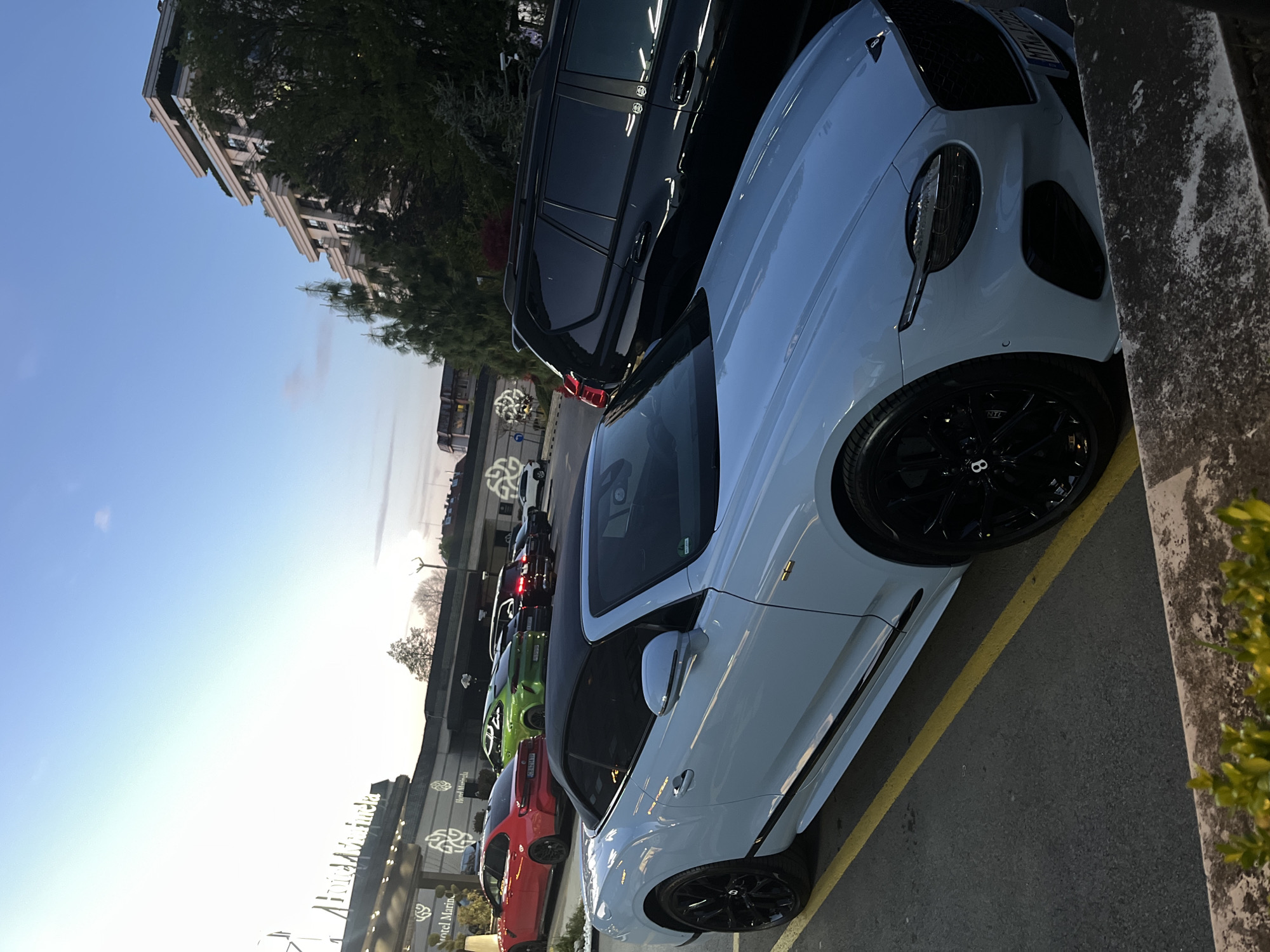 The image shows a row of parked luxury cars, including a white Bentley and a black SUV, in front of Hotel Marinela in Sofia, Bulgaria. Other vehicles are visible further down the row, including red and green cars. The sky is a light blue with the sun low on the horizon. A building is seen in the background on the left. The cars are parked in designated parking spots marked with yellow lines. A concrete wall with some vegetation is on the right.