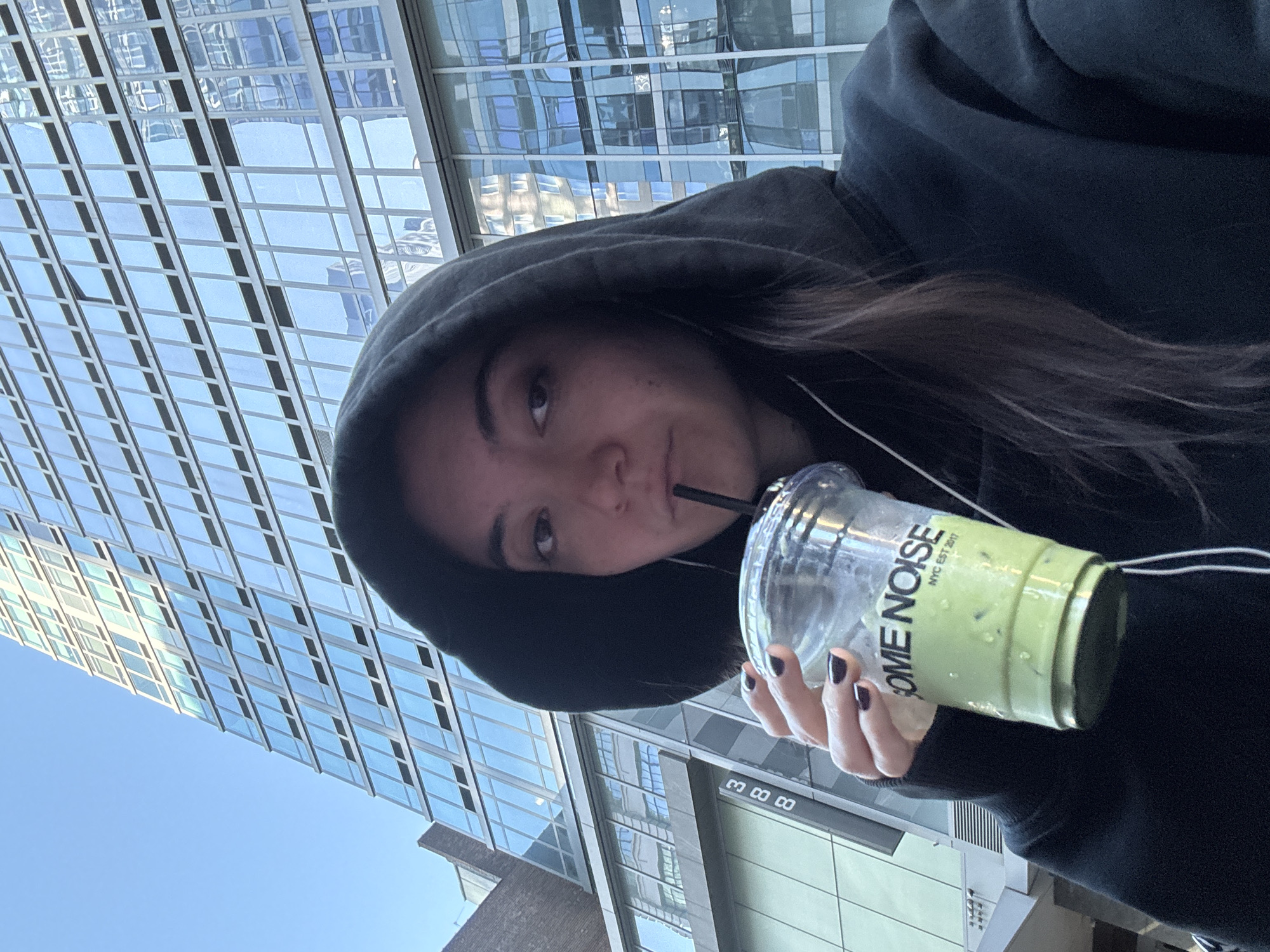 A woman wearing a black hoodie is holding a clear plastic cup with a green drink and a black straw, which she is sipping from. She has dark brown hair and dark nail polish. The cup has text that reads "SOME NOISE NYC EST 2017". The scene appears to be outdoors in an urban environment, with a modern glass building in the background, suggesting a city setting in New York, United States. The sky is blue, indicating it is daytime, and the lighting suggests it could be a sunny day. The woman is looking towards the camera, with a slight smile. The overall mood is casual and relaxed.