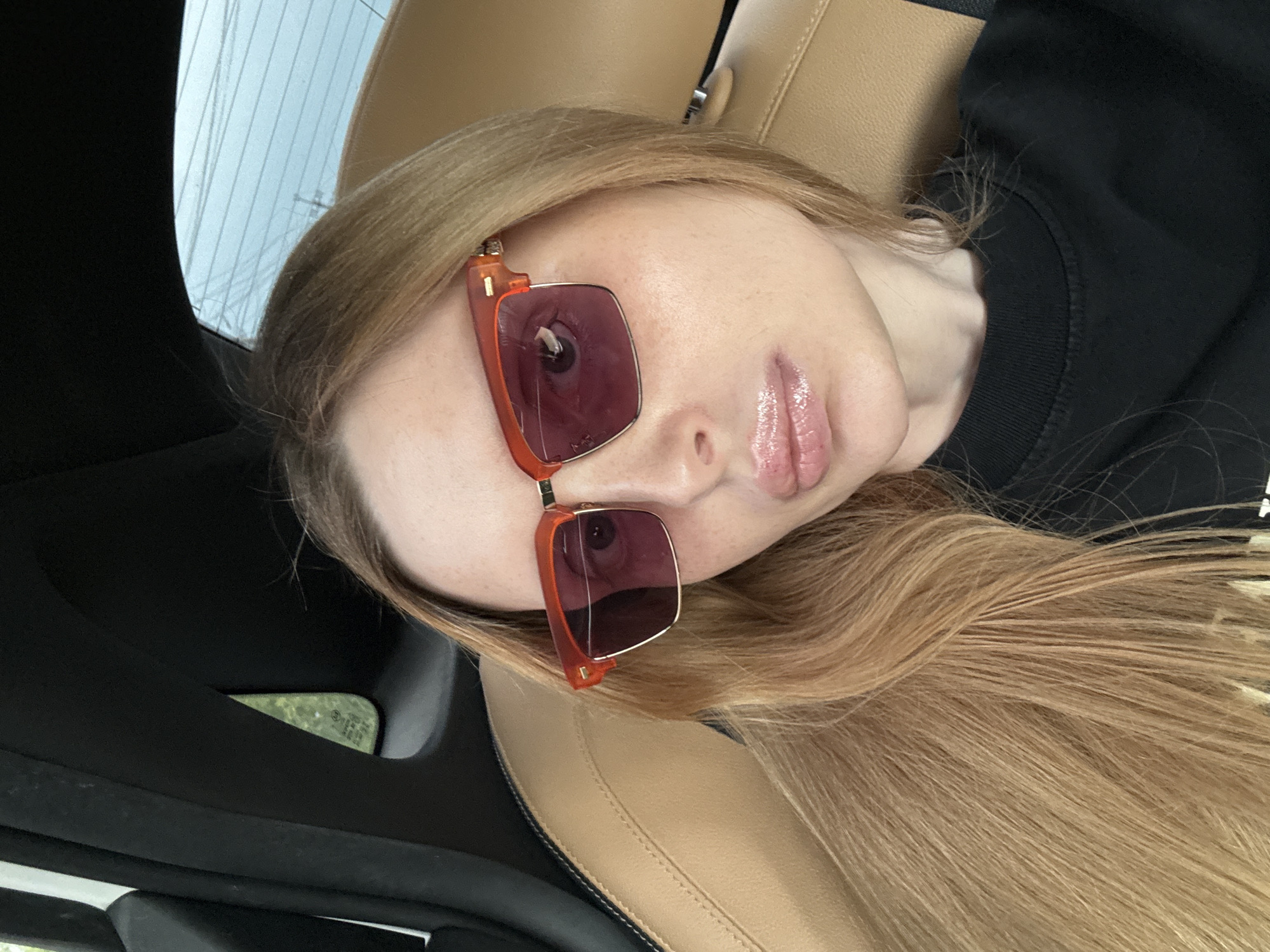 A close-up, eye-level, indoor selfie shows a fair-skinned woman with long, blonde hair, wearing large, translucent orange-framed sunglasses with pink lenses. She is seated in a car, with the leather seat visible in the foreground and the interior of the car behind her. The woman is looking directly at the camera, and her lips are slightly parted and glossy. Her facial features include freckles and a prominent cupid's bow. The background of the image is out of focus, with hints of green foliage seen through a car window.