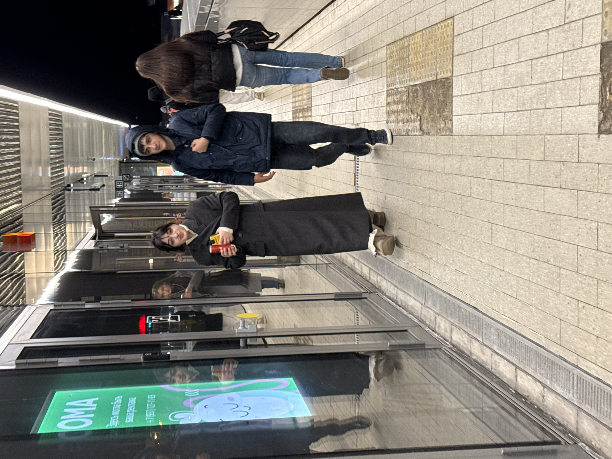 The image depicts a scene at a metro station in Khimki, Russia. Three people are standing on the platform. A woman is dressed in a long black coat and holding a red object. A man in a dark blue coat and a beanie is standing to her left. A woman wearing a jacket and jeans, with a backpack, is standing slightly behind them. The platform floor is paved with light gray rectangular stones and some sections with textured yellow stones. A metro train is visible through the station doors, displaying a brightly lit advertisement.