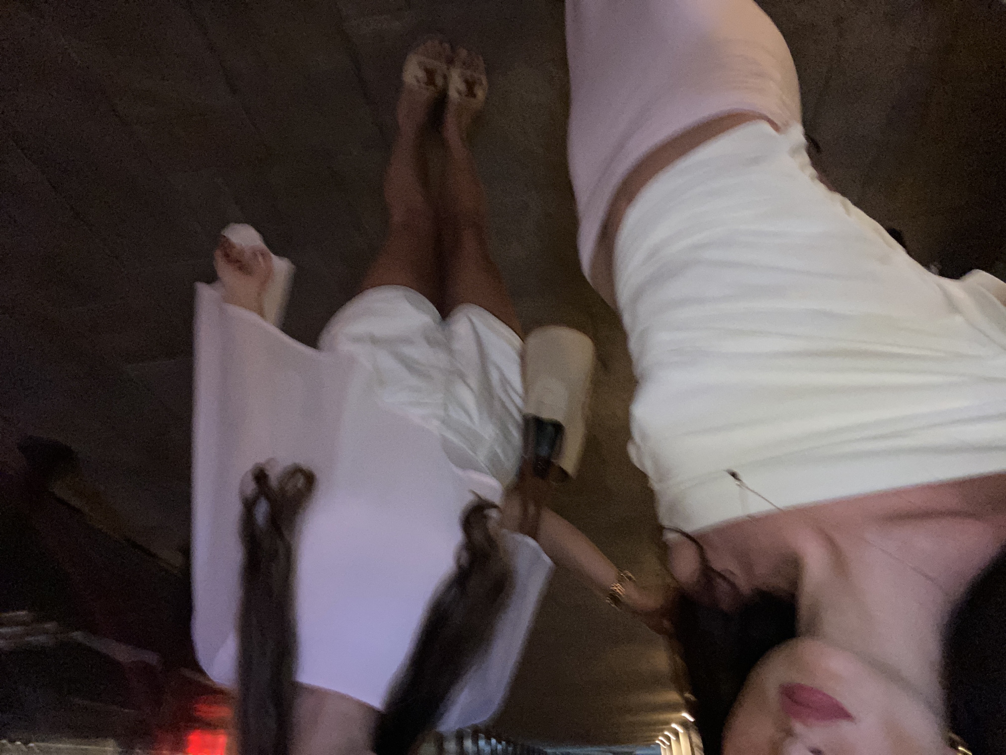 The image appears to be taken at night, possibly in Dubai, United Arab Emirates, based on the architecture visible in the background and overall impression. Two women are the primary subjects; one is wearing a light pink top and white bottom, and the other is wearing a white outfit. They seem to be in an outdoor setting with the city skyline faintly visible. The picture's orientation makes it difficult to clearly discern the environment or interactions between the subjects. The subjects are facing the camera with makeup visible on their faces. The scene is dark, with light coming from the background buildings.