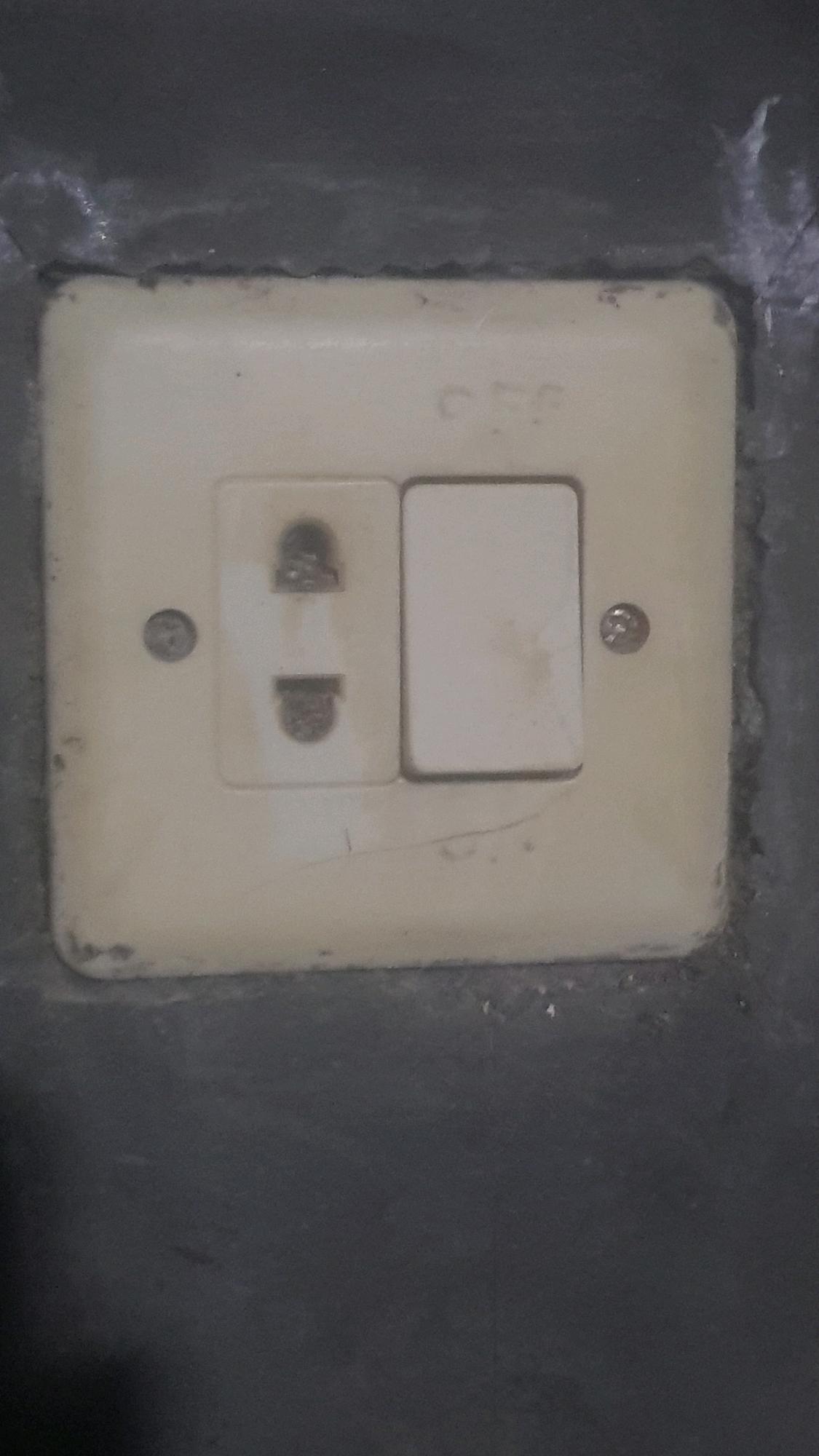 The image shows a close-up of an electrical outlet and light switch mounted on a dark gray wall. The outlet is cream-colored and has two rectangular slots. The light switch is also cream-colored and is located to the right of the outlet. The outlet and switch are set into a slightly larger cream-colored rectangular plate. There are screws visible on either side of the plate. The wall appears to be painted a dark gray and has a rough texture. The word "OFF" is faintly visible above the outlet and switch. The overall impression is of a functional, but somewhat aged, electrical fitting in a residential or commercial building. There are no people or any other objects visible in the image. This scene is located in Bakalan, Indonesia.