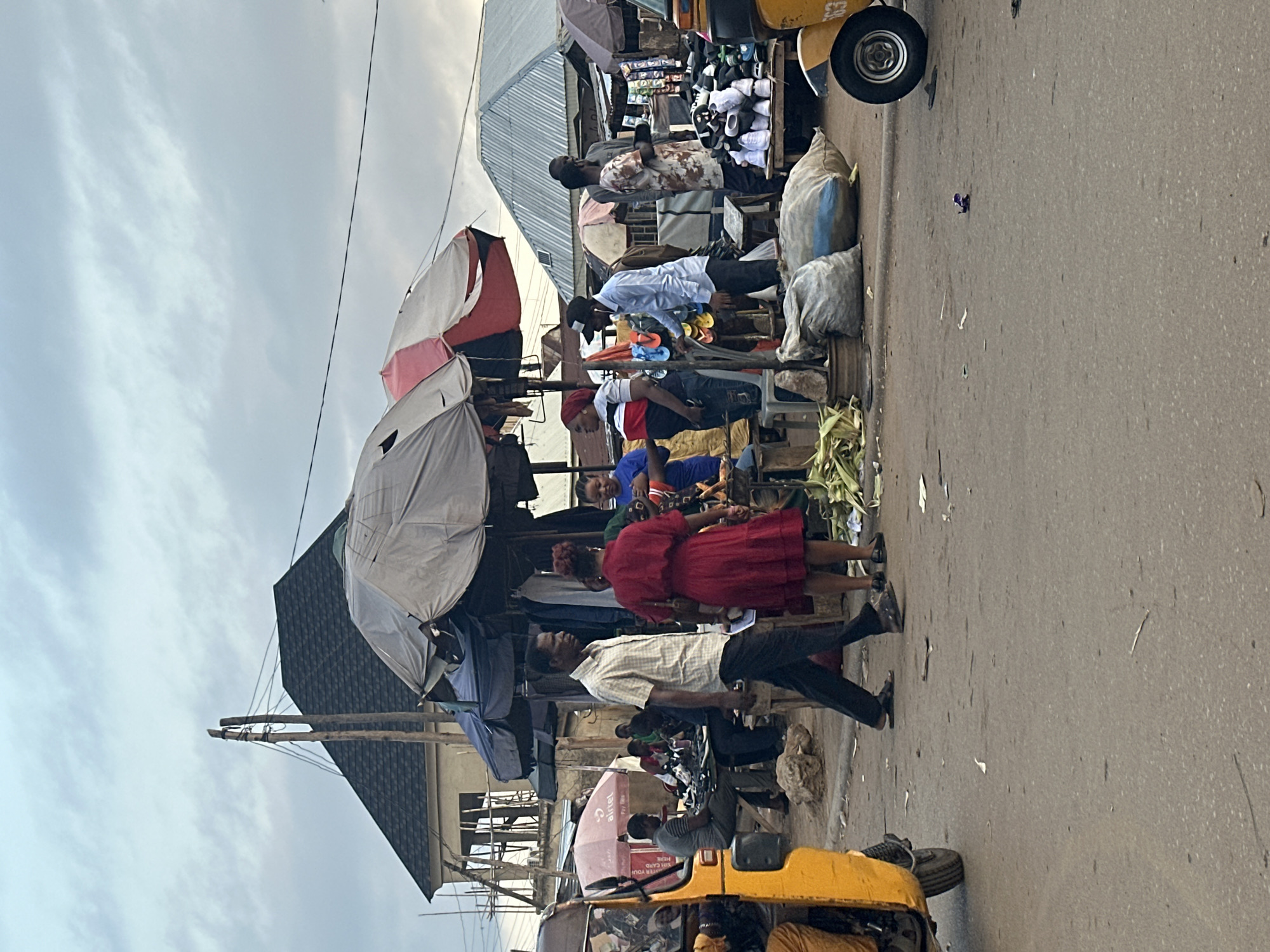 The image depicts a bustling street scene in Abuja, Nigeria, likely a market area. The orientation of the image is sideways, with the sky appearing on the left and the ground on the right. In the foreground, a yellow motorized tricycle, commonly known as a "Keke," is partially visible. The road surface is asphalt, with some debris scattered on it.
The middle ground of the image is filled with market stalls and people. Several market stalls are set up with awnings, some covered with white and pink fabric, and others with umbrellas. Goods are displayed, including what appear to be shoes and clothing. Several people are seen in the market, interacting with each other and attending to their businesses. A woman in a red dress is a prominent figure, as is a man in a checkered shirt and dark pants. Another man is leaning over a stall, possibly a vendor. Some people are seated, while others are standing. There are also large sacks, likely containing goods, stacked near the stalls. The overall atmosphere suggests a busy commercial environment.
In the background, more market structures and people can be discerned. The sky is overcast, suggesting it might be a cloudy day. The lighting appears diffused, indicating it could be either early morning or late afternoon. The presence of the Keke and the style of the market stalls are strong indicators of the location in Nigeria. The image captures a slice of everyday life in a busy urban setting. There is text visible on a pink umbrella which reads "Airtel".