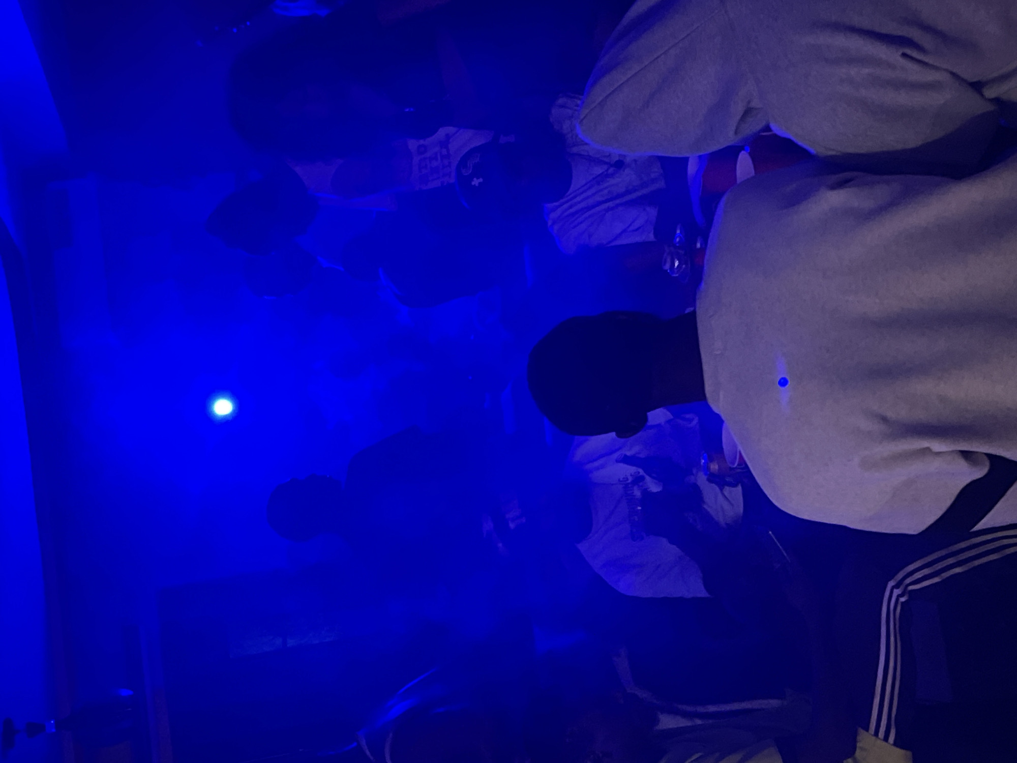 The image depicts a dimly lit indoor gathering, predominantly illuminated by intense blue lighting, with some visible haze or smoke. Several individuals are present, their faces largely obscured by shadows and the ambient light. In the foreground, a person wearing a light-colored crewneck sweatshirt is facing away from the camera, with a subtle blue light reflecting on their back. To their right, another individual in a grey hooded sweatshirt is also visible from the back.
In the mid-ground, several other people are gathered, some appearing to be seated. One individual in the center is wearing a white t-shirt and a baseball cap, with hands clasped together. Another person to their left is wearing a white t-shirt with a graphic print. Objects such as cups and what appear to be cans are visible on a surface in the mid-ground, suggesting a social event.
The overall environment appears to be a somewhat enclosed space, possibly a bar, club, or private party venue, given the lighting and gathering of people. There is no specific visual information in the image to determine its location within Durumi, Nigeria.
