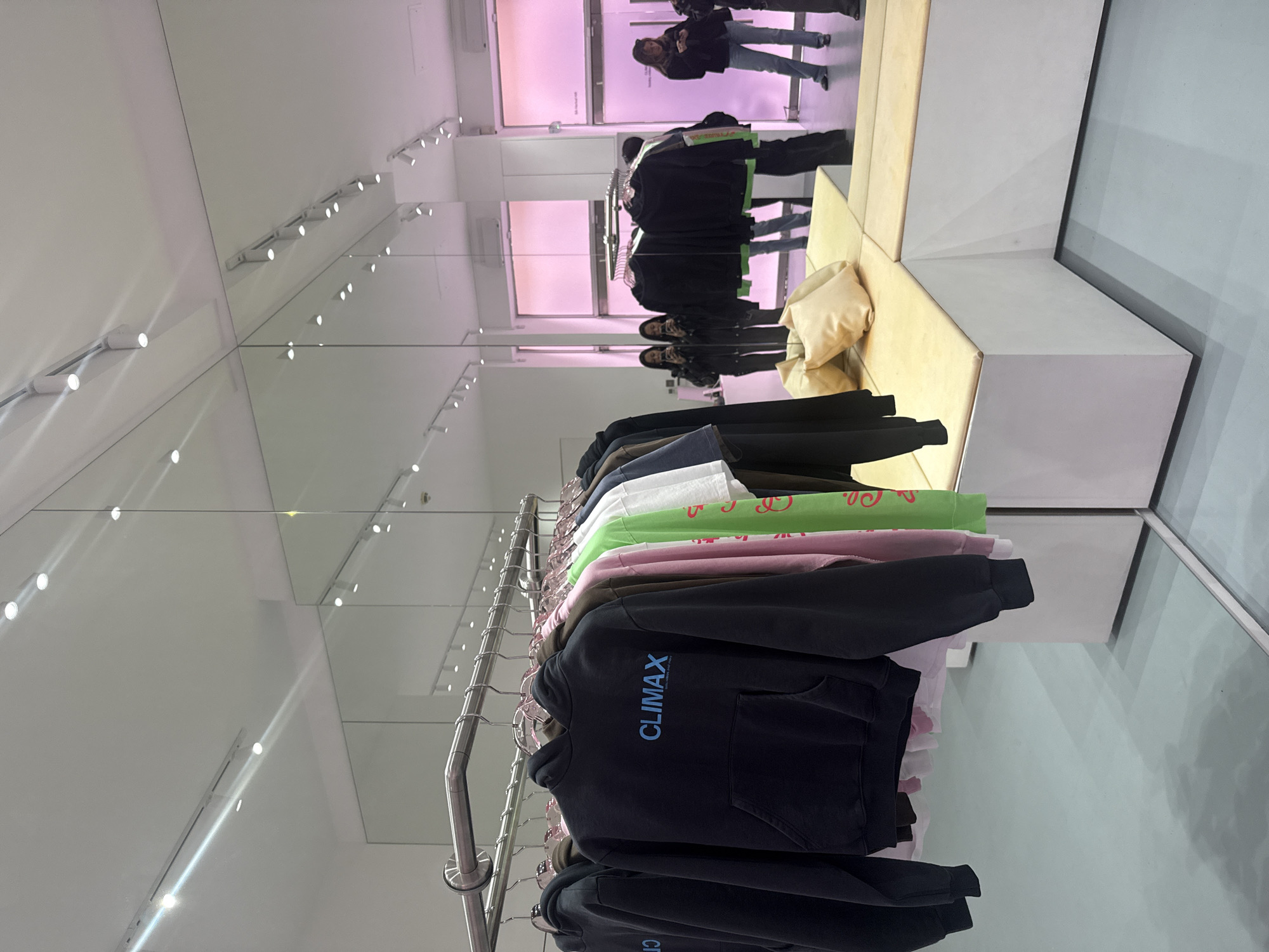 This image depicts a modern retail space or showroom, likely in London, United Kingdom, characterized by extensive use of mirrors. The central focus is a metal clothing rack displaying a collection of hooded sweatshirts (hoodies) and t-shirts in various colors, including black, brown, pink, light green, and white. The prominent hoodie in the foreground is dark blue or black, featuring the brand name "CLIMAX" printed in light blue capital letters on the chest.
The walls are predominantly mirrored, creating multiple reflections of the merchandise, the interior architecture, and people within the space. Bright track lighting is installed on the white ceiling, and its reflections contribute significantly to the bright and expansive feel of the room.
In the background, beyond the clothing rack, reflections reveal several individuals. One person, wearing a dark jacket and jeans, stands facing a section of the store bathed in a distinctive soft pink ambient light. Further reflections show other individuals, appearing to be browsing or waiting. A minimalist seating arrangement, comprising light-colored, possibly upholstered, geometric benches, is visible on the right side of the image, with a beige cushion or bag resting on it. The floor is a light, reflective surface, mirroring parts of the display. The atmosphere is sleek and contemporary. The specific time of day is not discernible, but the setting is clearly indoors during operational hours.