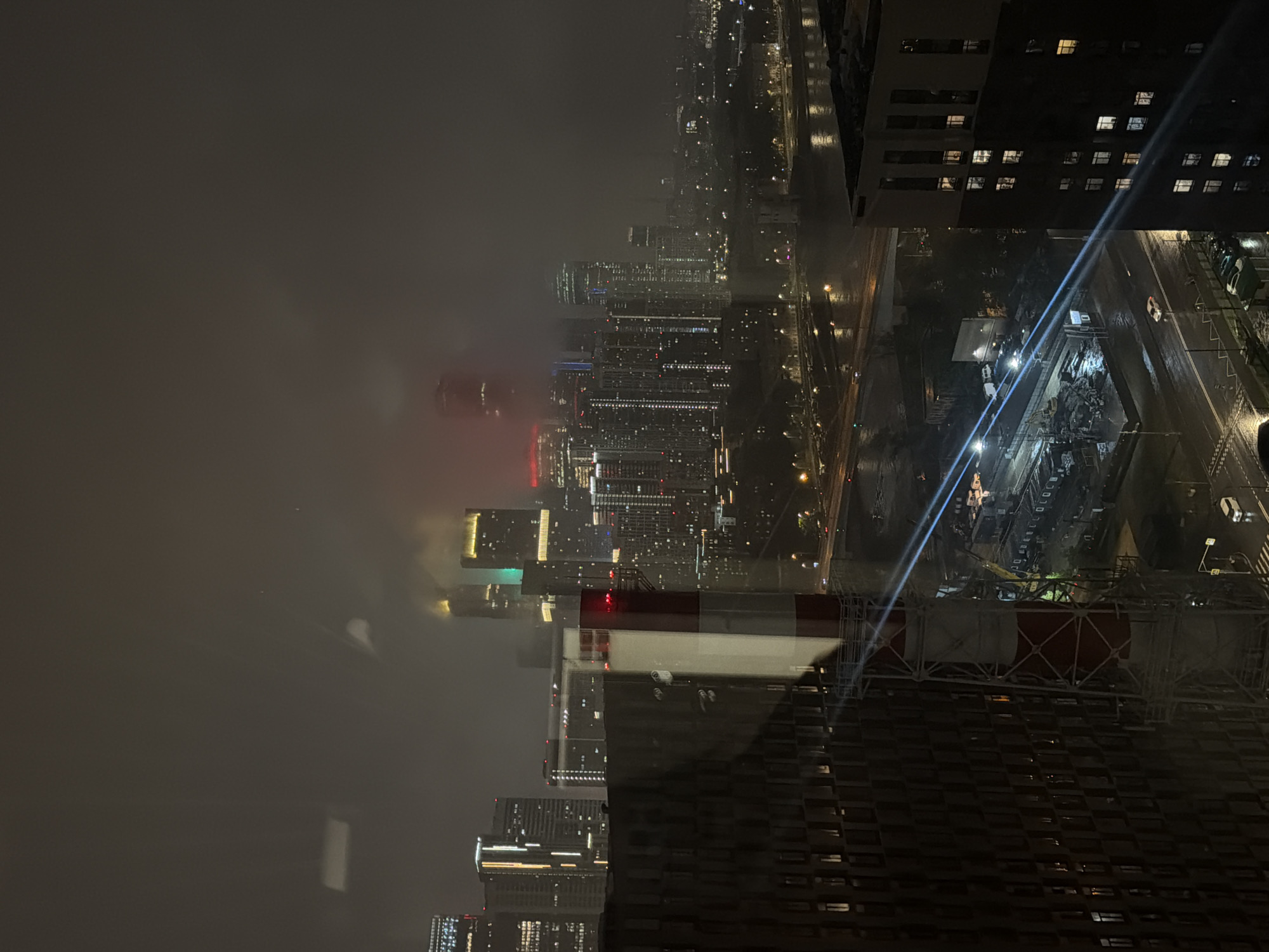 This image captures a nocturnal, atmospheric view of a modern city skyline, likely Khimki, Russia, shrouded in mist or fog. In the foreground, a large, industrial-looking structure, possibly a chimney or tower with red and white striped markings, dominates the left side. It is supported by a complex metal framework. To the left of this structure, a dark, multi-story building with numerous illuminated windows is visible, suggesting residential or commercial use.
Beyond the foreground elements, a dense cityscape unfolds, characterized by numerous tall buildings of varying designs and heights, illuminated by a multitude of lights. Some buildings display prominent vertical lighting. The sky above is dark and obscured by atmospheric conditions, with faint glows suggesting artificial light pollution. A bridge with traffic lights and vehicle lights spans across a body of water in the mid-ground. Below the bridge, streetlights illuminate a wet road surface, indicating recent rain. Various vehicles, including cars and a van, are visible on the roads. In the distance, the city lights blend into the fog, creating a hazy and somewhat ethereal panorama. The overall impression is one of urban density at night, with a strong sense of depth and a slightly subdued color palette due to the low light and atmospheric conditions.