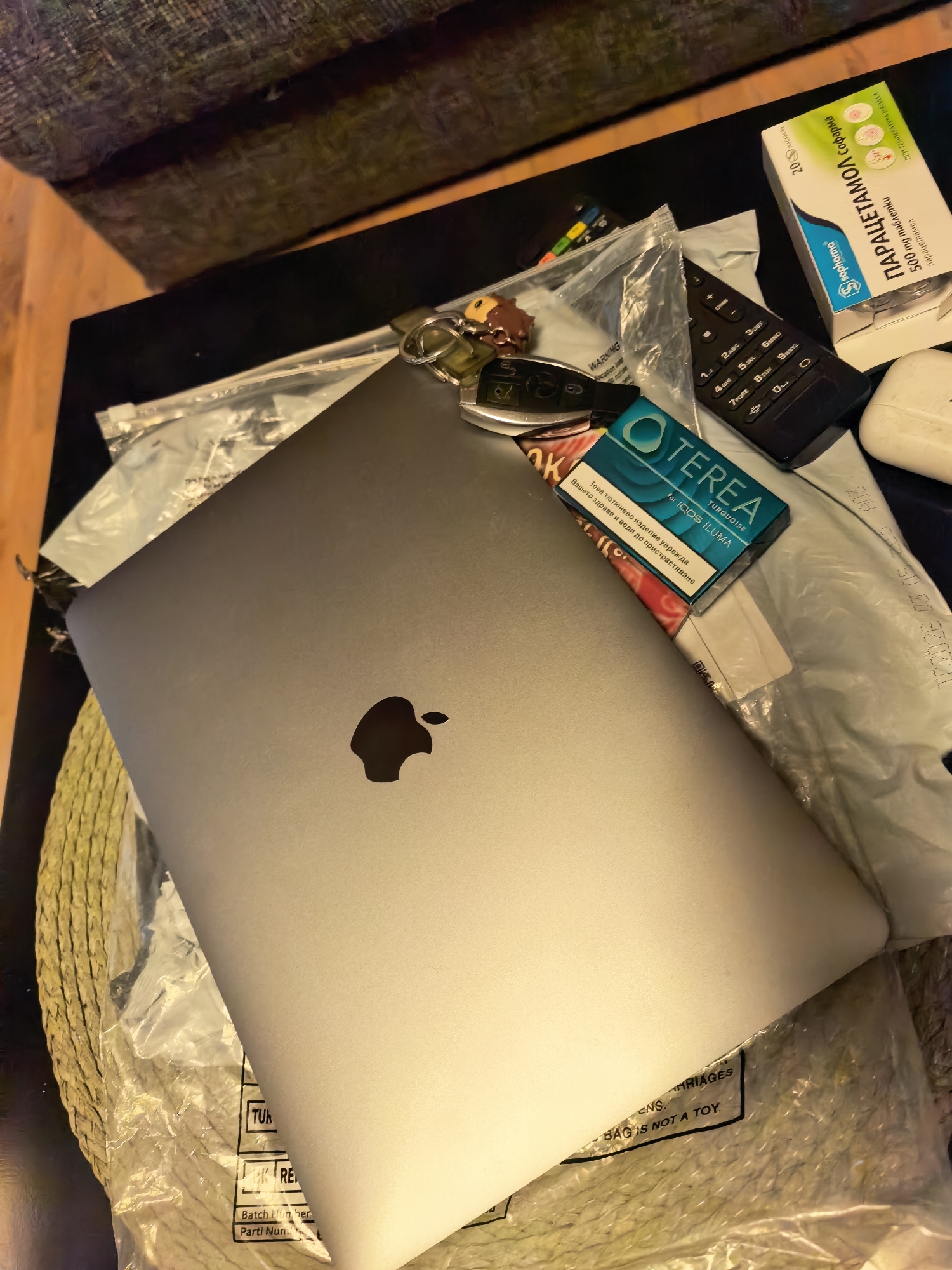 The image shows a cluttered tabletop in Sofia, Bulgaria. At the center lies a closed silver MacBook. On top of the laptop are a set of keys, a black car key fob, and a pack of "TEREA Turquoise for IQOS ILUMA" tobacco sticks. To the right of the laptop is a black remote control, a white earbud case, and a box of "Paracetamol Sopharma 500mg" tablets. The items are resting on a partially visible woven placemat that appears to be inside a clear plastic bag. The bag has a warning label "BAG IS NOT A TOY." The table itself appears to be black. The edge of a chair or sofa is visible in the background. The presence of Bulgarian text on the TEREA and Paracetamol packaging indicates the location as Sofia, Bulgaria.