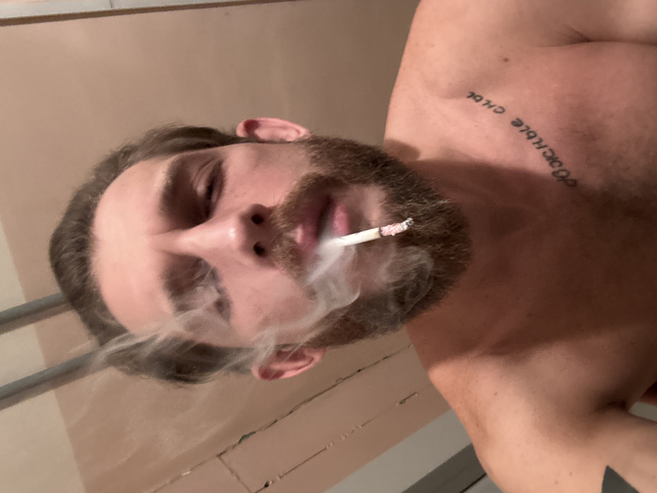 A shirtless man with a beard is holding a lit cigarette in his mouth, exhaling smoke. He appears to be indoors, the room lit with a dim, warm light. A tattoo is visible on his upper chest. The camera angle is low, looking up at the man's face. The background is plain and indistinct. The man is the primary focus, with the cigarette smoke adding a dynamic element to the composition.