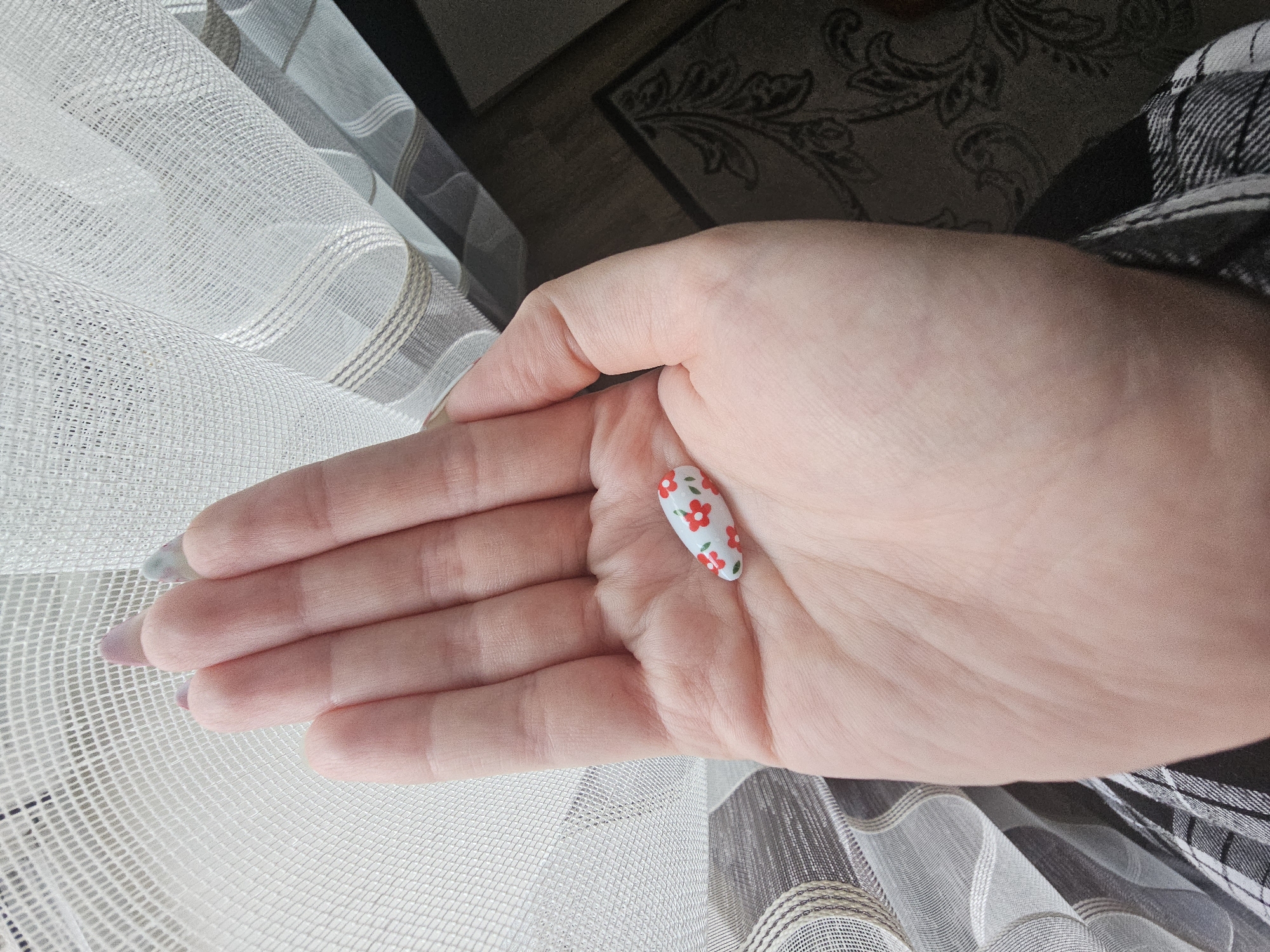 A person's left palm is holding a small, oval-shaped object that appears to be a fake fingernail. The fingernail is white with a red floral pattern. The person's hand is resting against a sheer, white, patterned curtain. The background is out of focus and appears to be a dark rug with a floral design. The image was taken indoors. There is no information about the city of Kamen-na-Obi, Russia included in the image.