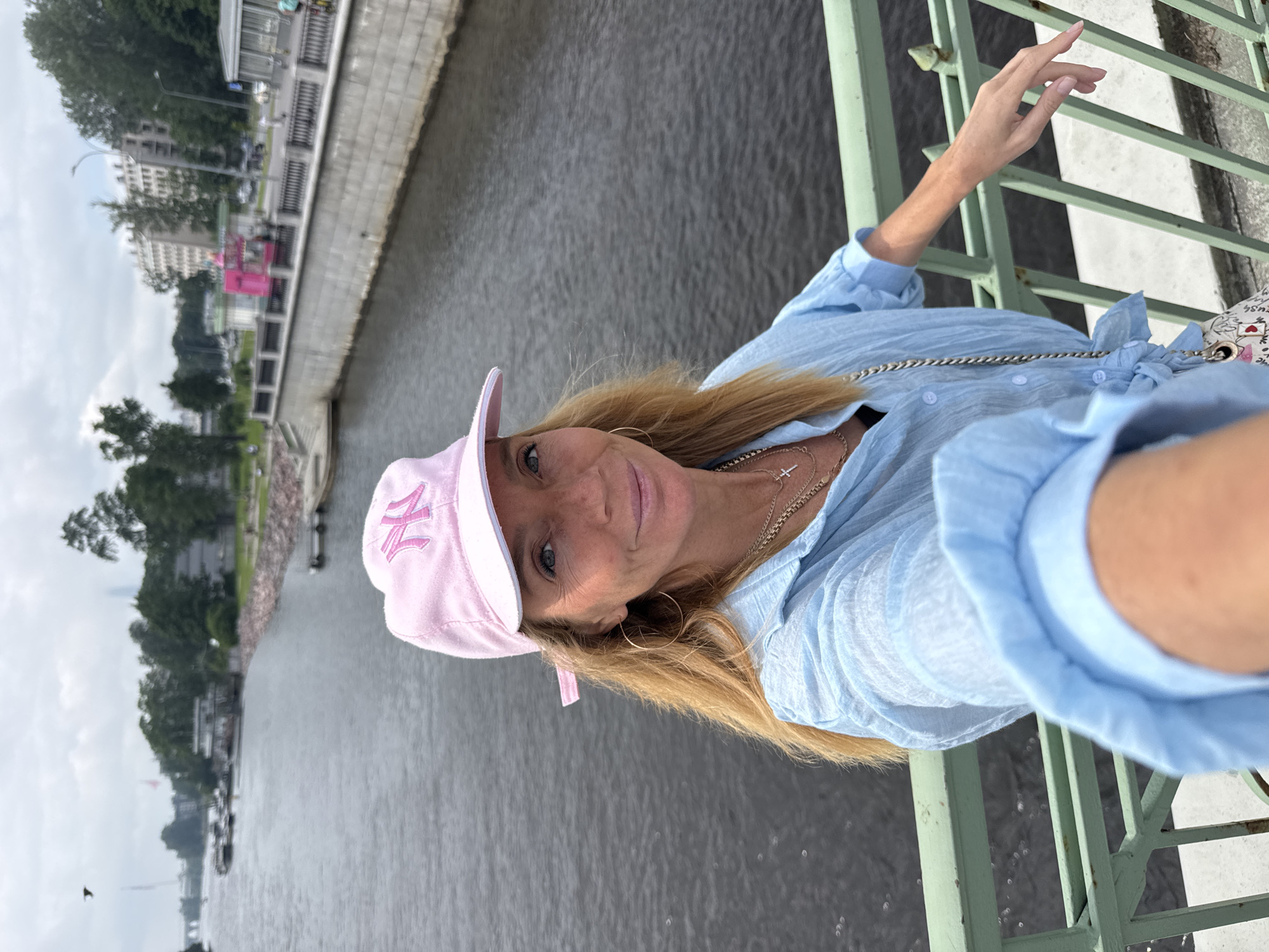 A woman wearing a pink baseball cap and a light blue, short-sleeved shirt is taking a selfie on a bridge over a body of water. She has long blonde hair and is wearing hoop earrings and several necklaces. In the background, there is a river, buildings, trees, and a cloudy sky. The bridge has green railings. This appears to be the Neva River in Saint Petersburg, Russia.