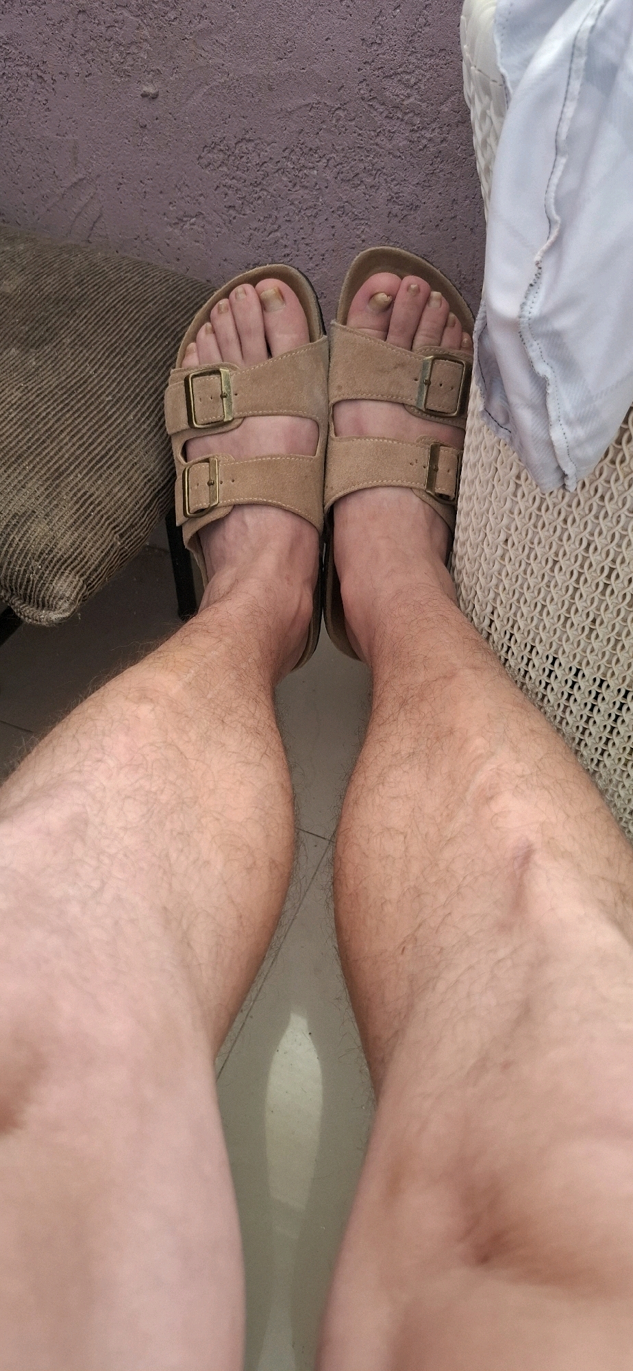 A pair of lightly tanned legs wearing tan Birkenstock sandals are shown from a high-angle perspective. The feet are positioned side-by-side, with the toes pointing slightly outwards. The sandals are open-toed, with two adjustable straps across the top of each foot. The left sandal's straps are buckled, and the right sandal's straps appear to be buckled as well. The toenails are visible and appear to be unpolished and slightly discolored. The legs are covered in light hair. The background consists of a textured, light purple wall on the left, a textured brown object on the far left, and a white woven textured surface on the right. The floor is a light gray tile. The overall setting appears to be indoors. The image's context of being in Yerocham, Israel, is a given environmental detail.