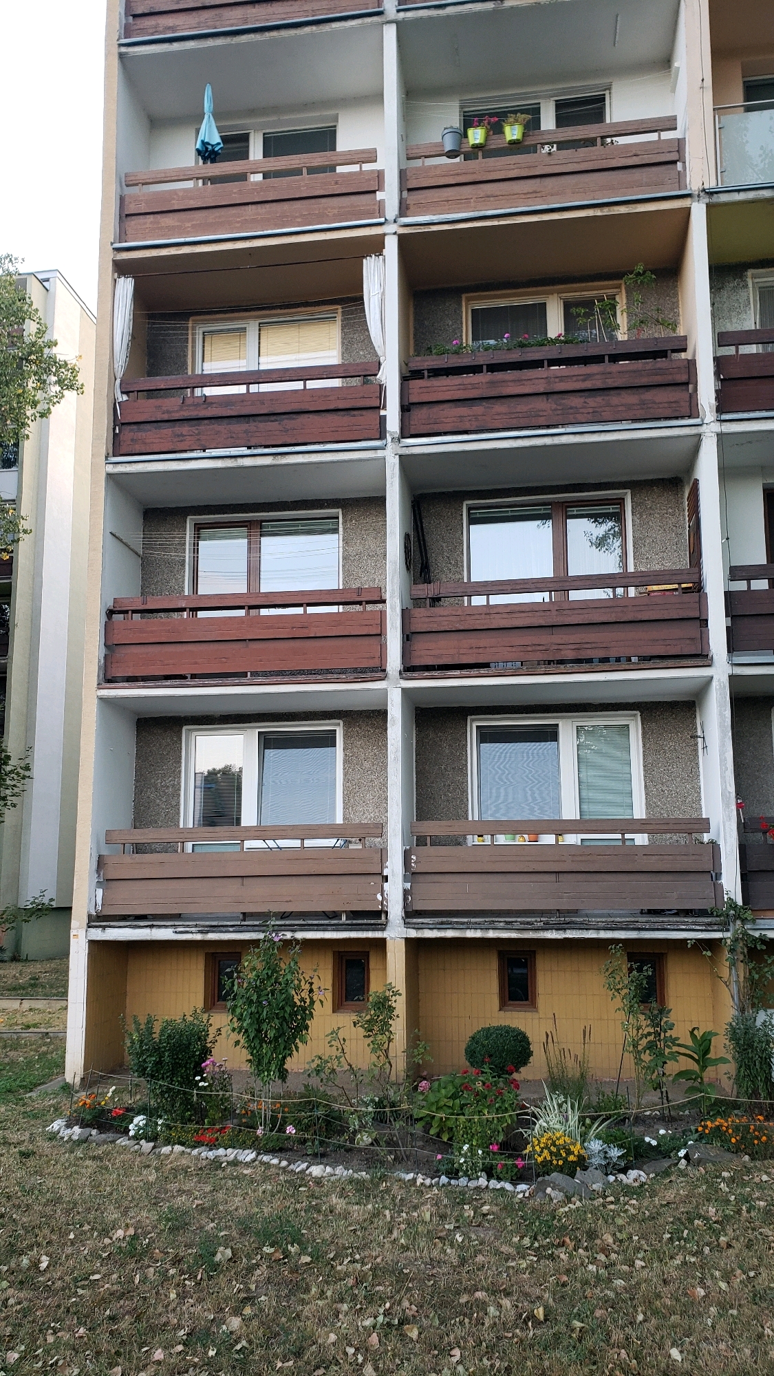 The image shows a multi-story apartment building with balconies on each floor. The building appears to be located in a residential area, possibly in the city of Prešov, Slovakia, given the context provided. The balconies are furnished with wooden railings and some have plants. The lower level of the building features a small garden area with various flowers and shrubs, enclosed by a low fence. The overall scene depicts an exterior view of a residential building on a cloudy day. There are no people visible in the image.
