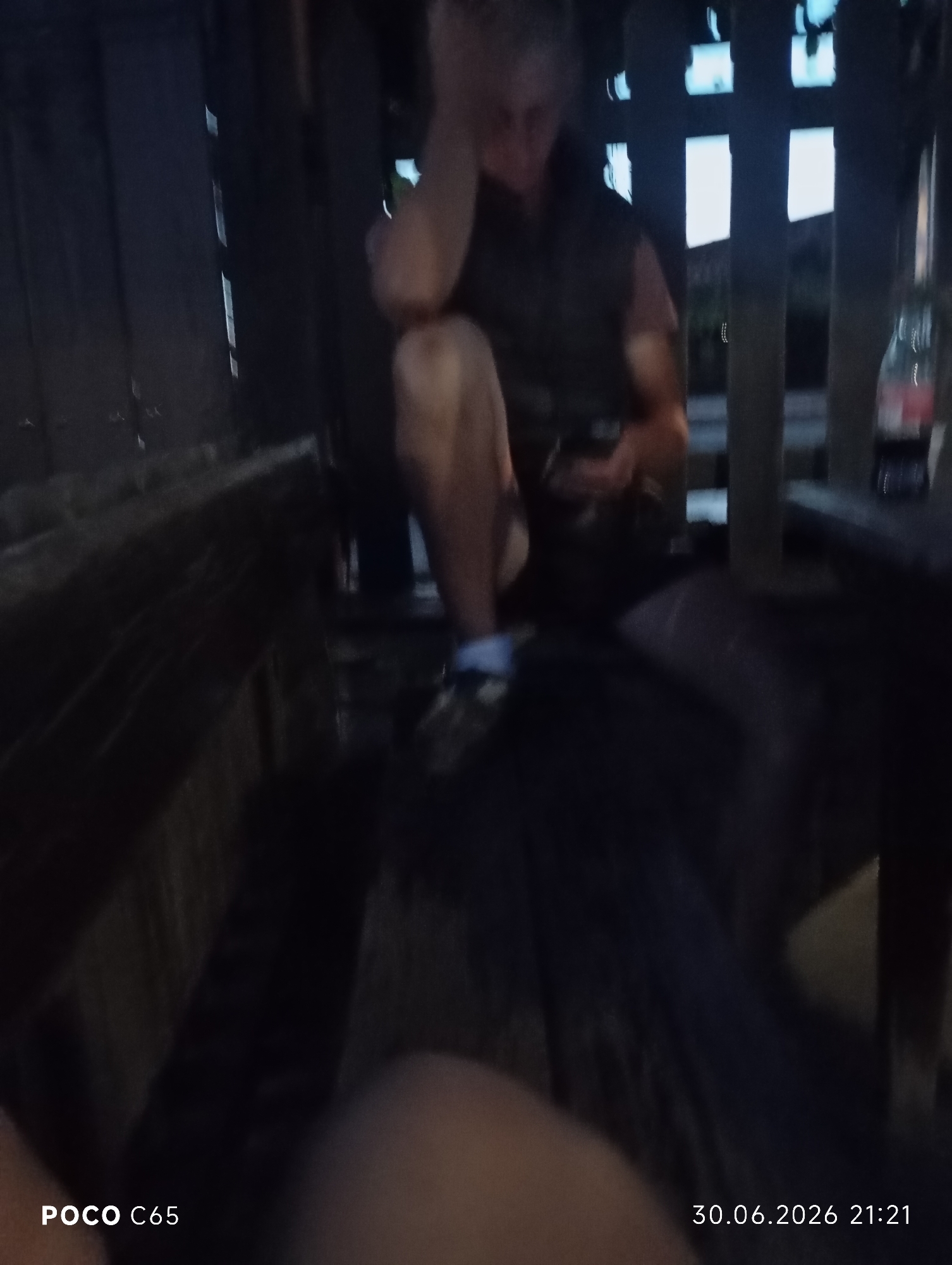 The image depicts a dimly lit, outdoor scene, likely in Kozichino, Bulgaria, at night. The central subject is a person, appearing to be male, sitting down with their hand on their head, suggesting a relaxed or contemplative pose. They are wearing dark clothing and what appear to be light-colored shoes with dark accents. The setting seems to be a rustic wooden structure, possibly a deck or a makeshift seating area, with dark wooden planks visible throughout the frame.
In the background, through gaps in the wooden structure, glimpses of illuminated rectangular shapes can be seen, possibly windows or lights from a building. A red bottle, likely a beverage, rests on a surface to the right of the central figure. The overall ambiance is dark, suggesting nighttime, and there are no clear indicators of weather conditions.
The image also contains visible text at the bottom left and right corners. The bottom left displays "POCO C65", indicating the model of the phone used to capture the image. The bottom right shows a date and time stamp: "30.06.2026 21:21".