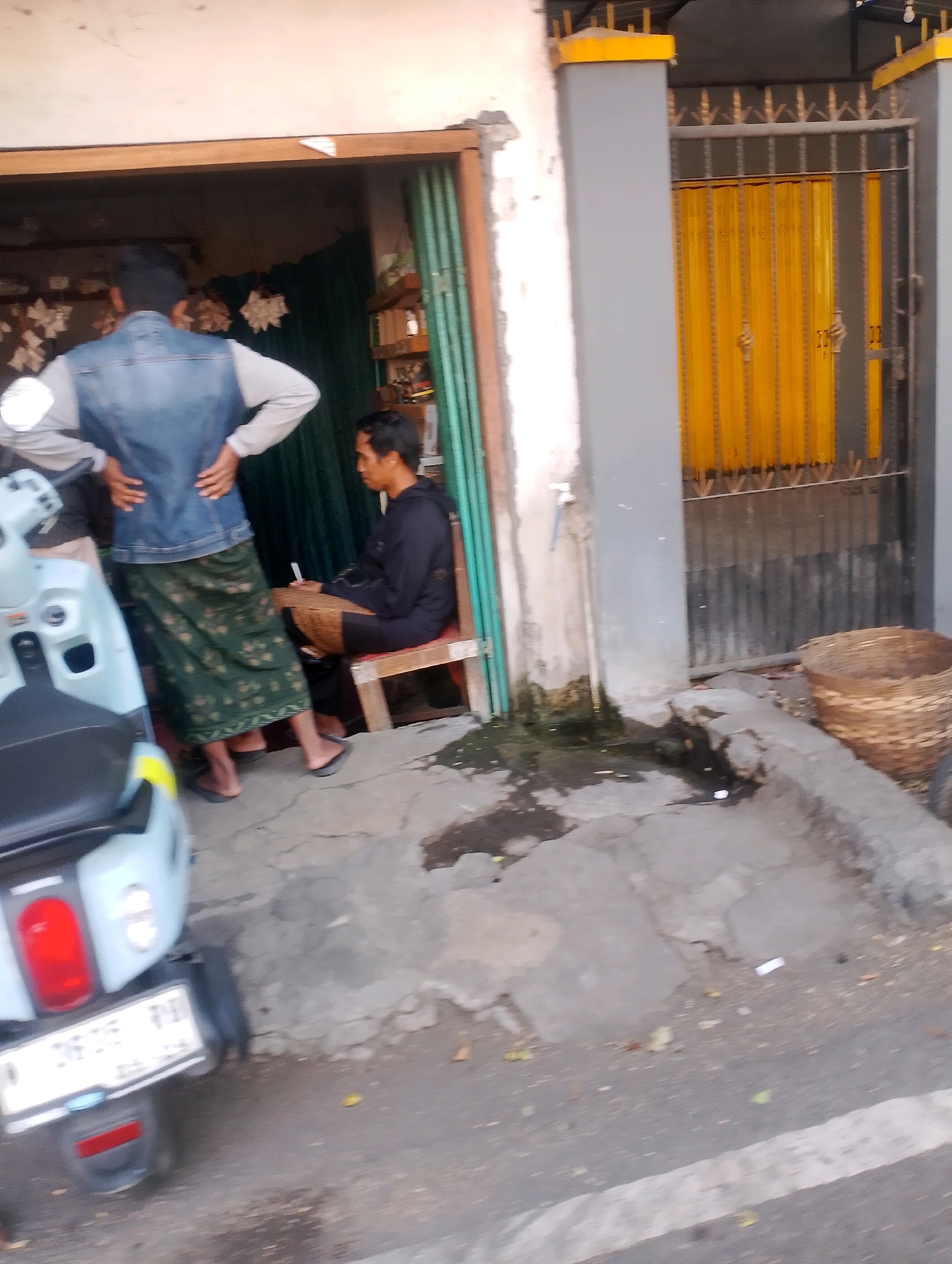 The image depicts an outdoor scene in Probolinggo, Indonesia, likely on a street in front of a building. In the foreground, the rear of a light blue scooter is visible, featuring a prominent red taillight and a white license plate. The license plate displays characters that are illegible due to the image resolution.
To the left of the scooter, a person is partially visible from the back, wearing a denim vest over a light-colored long-sleeved shirt and a green sarong with a floral pattern. Their hands are on their hips.
In the middle ground, a man is seated on a simple wooden chair, wearing a dark long-sleeved shirt and a brown sarong. He is holding a cigarette or similar object. He is positioned in what appears to be the entrance of a shop, indicated by green folding doors and an open doorway. Inside the shop, glimpses of shelves and possibly goods can be seen, along with a green curtain.
To the right of the shop entrance, a yellow corrugated metal door is visible behind a metal gate with decorative finials. A large, woven basket sits on the ground to the right of the entrance, near a concrete gutter. The ground surface is a mix of cracked concrete and asphalt. The overall environment suggests a commercial or residential area.
