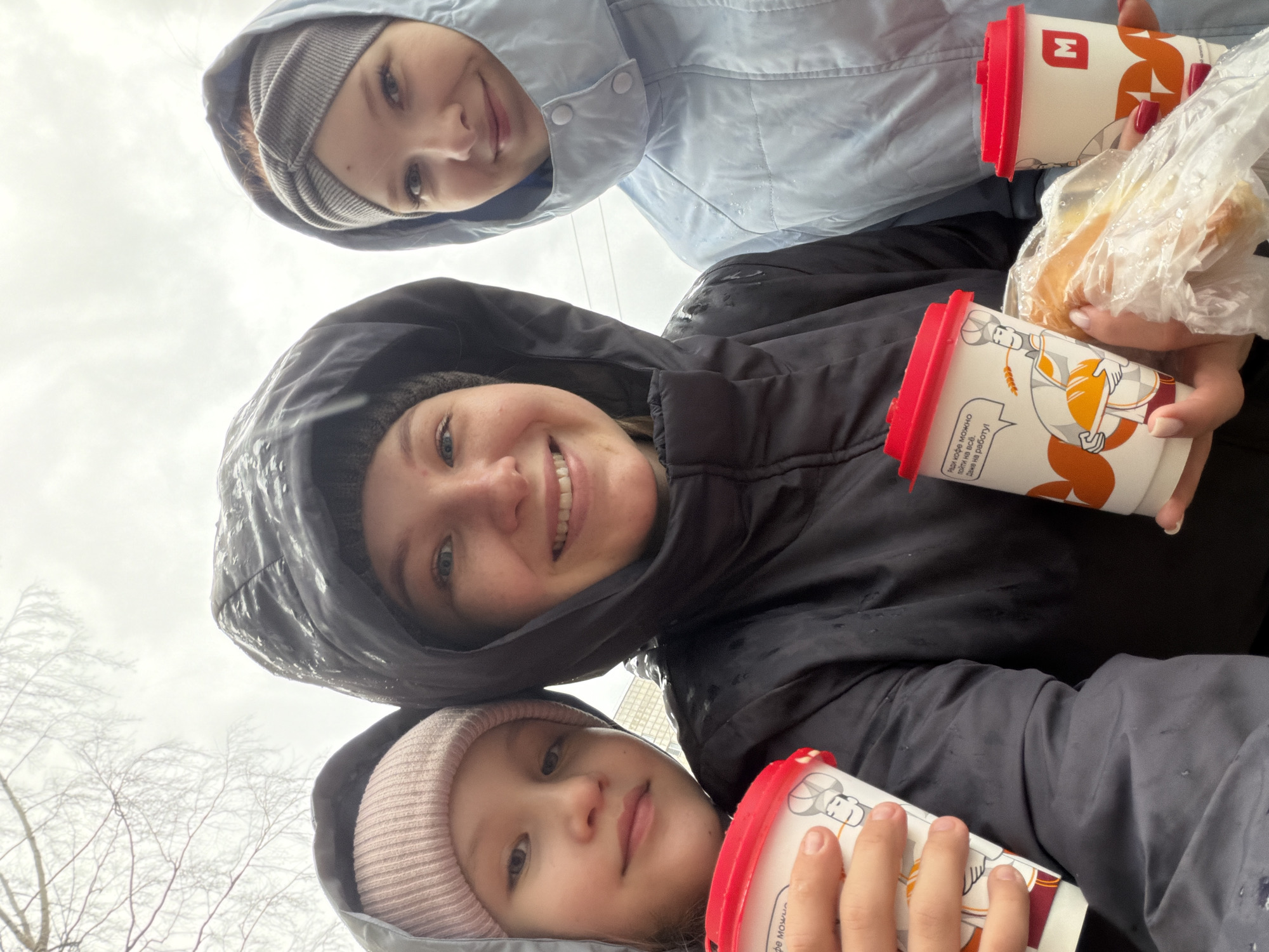 Three people, likely family members, are clustered together outdoors, facing downwards towards the camera. The person in the middle is a woman, who is smiling. The woman has two young girls next to her.
They are all wearing hooded jackets and hats. The two girls are wearing beanies under their hoods. They appear to be holding drinks, in white cups with red lids, from a chain food store called "М." They also are holding a plastic bag with baked items, likely from the same establishment.
Based on the presence of the chain food store "М" in the cup, along with Russian text on the cup, this image was taken in the city of Chaikovsky, Russia. The background is of bare trees against an overcast sky, indicating a cold season. The overall scene suggests a casual outing, possibly buying something to eat. The jackets look like they have water on them, which would further indicate that it is a rainy day.
