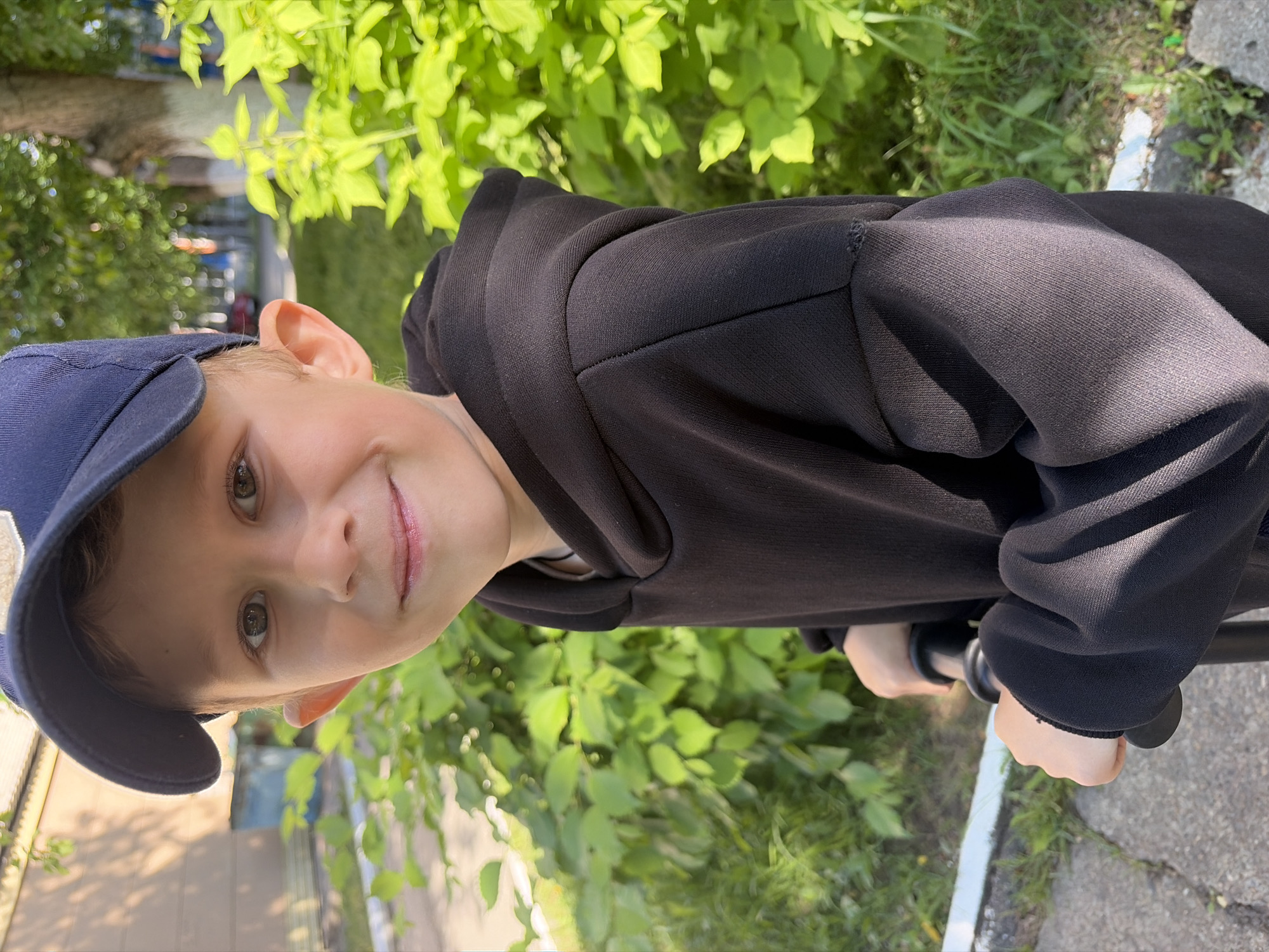 A child wearing a blue baseball cap and a black hoodie is positioned in front of a lush green bush and a section of sidewalk. The child's hands are visible, gripping what appears to be handlebars. The image is taken outdoors on a bright day. The specific location is Chaikovsky, Russia.