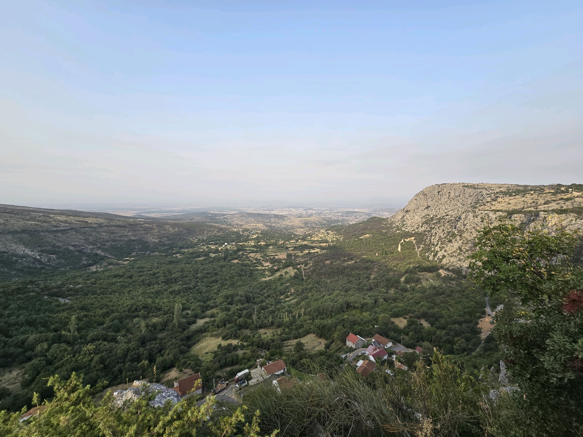 An elevated panoramic view looks across a valley towards a distant city under a pale blue sky. The foreground is dominated by dense green foliage and rocky outcrops. In the midground, a cluster of houses with red tile roofs are visible, situated along a winding road. This settlement is nestled within a valley covered by a thick expanse of green trees. Further in the distance, the valley opens up to reveal a more spread-out urban area with buildings and infrastructure. A prominent, rugged, light-colored rocky hill rises on the right side of the image, contrasting with the green landscape. The scene appears to be a rural or semi-rural area on the outskirts of a city, possibly Gornja Vrbica, Montenegro, given its hilly terrain and proximity to a larger settlement.
