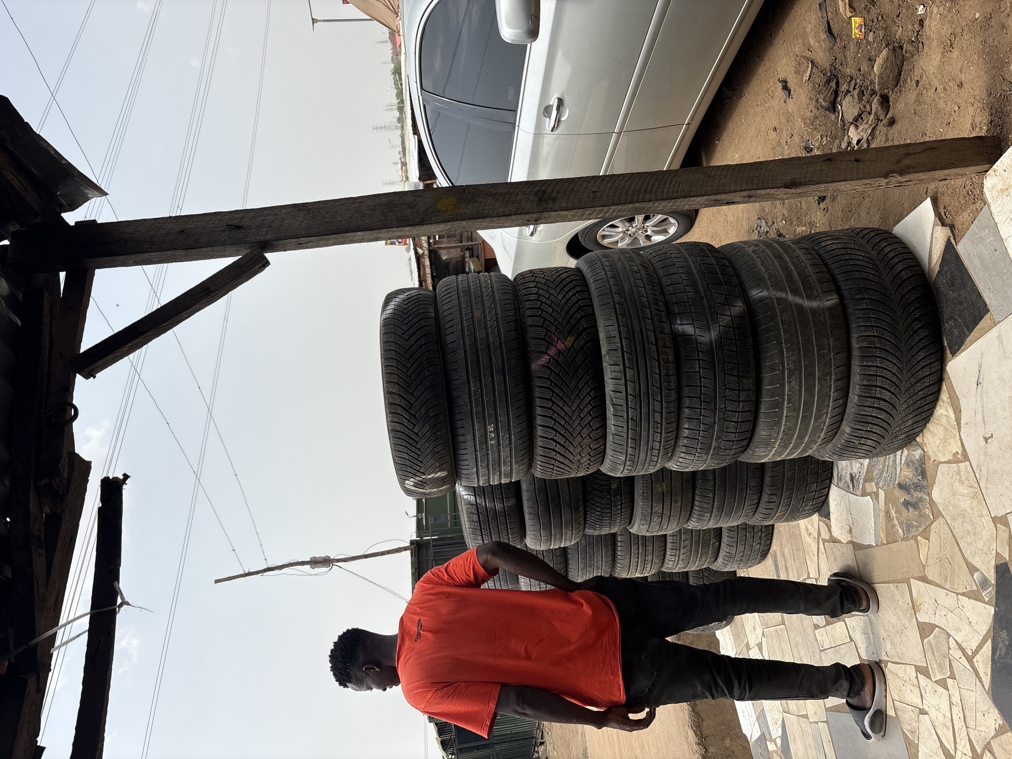 The image depicts a scene in Lugbe, Nigeria, showing a man standing beside a stack of tires. The tires are piled in a single column, approximately ten high, and are located near a white car. The man is wearing an orange t-shirt and black pants. The setting appears to be an outdoor tire shop or storage area. The ground is covered with dirt and broken concrete tiles. An overhead wooden structure provides partial shade. Power lines are visible in the background.