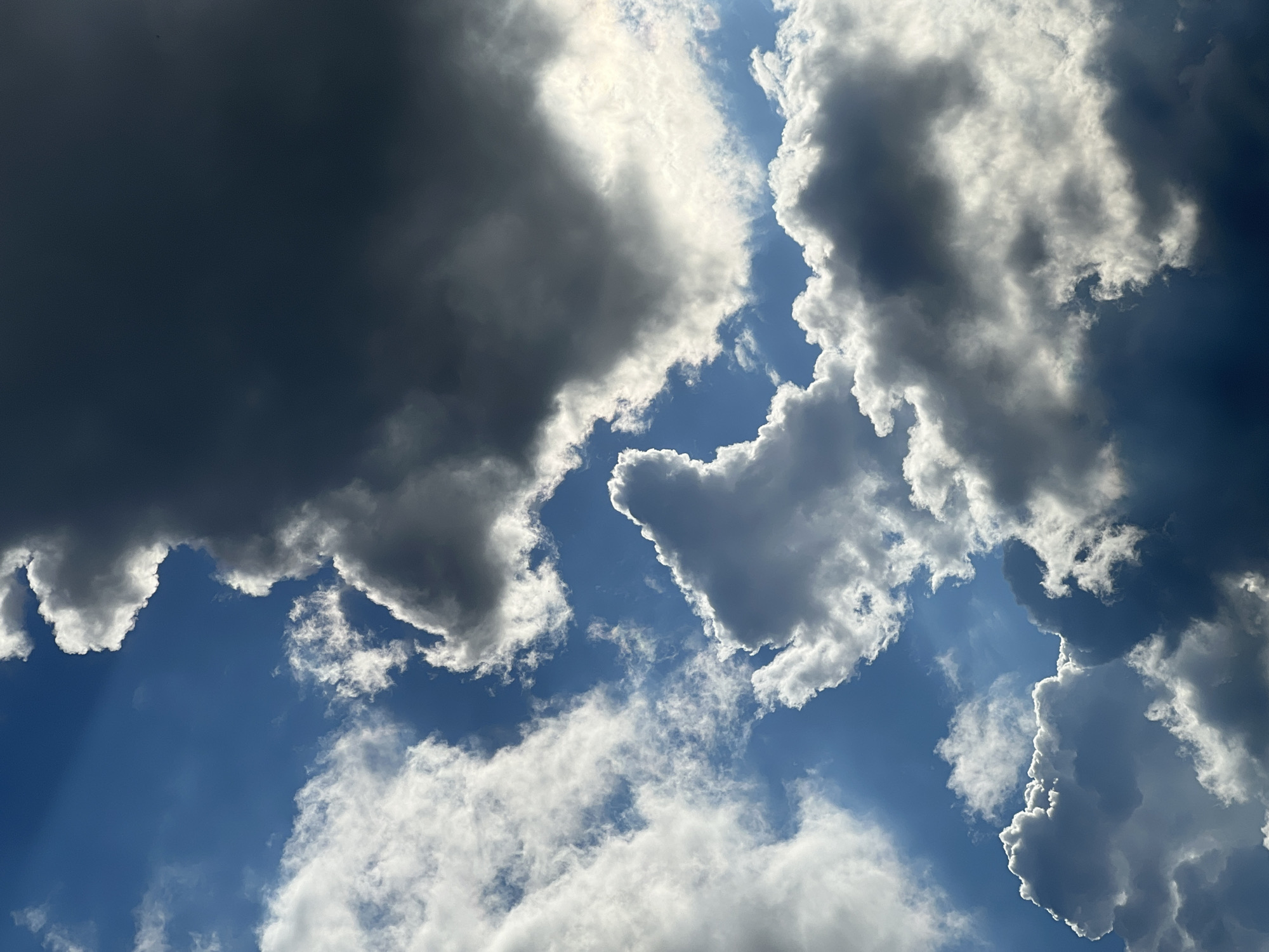 The image is an outdoor, low-angle shot of a sky filled with dynamic clouds against a bright blue sky. Sunlight pierces through some of the clouds, creating dramatic shafts of light. The clouds vary in density and color, ranging from bright white, illuminated by the sun, to dark gray, suggesting potential storm activity or shadow. There are no people, structures, or terrestrial elements visible in the frame, only the sky and clouds. The scene is captured without any discernible location markers specific to Warsaw, Poland.