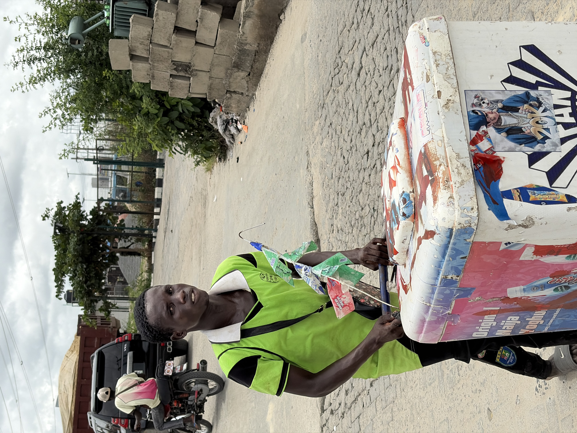 In Igboefon, Nigeria, a dark-skinned man in a bright green shirt with black trim stands on a paved street. He holds a collection of colorful pinwheels. Next to him is a weathered cart adorned with peeling posters and stickers, including one featuring an illustration of a rapper. A black motorcycle with a passenger is parked behind him. In the background, there is a stack of concrete blocks, foliage, and a glimpse of buildings under a cloudy sky.