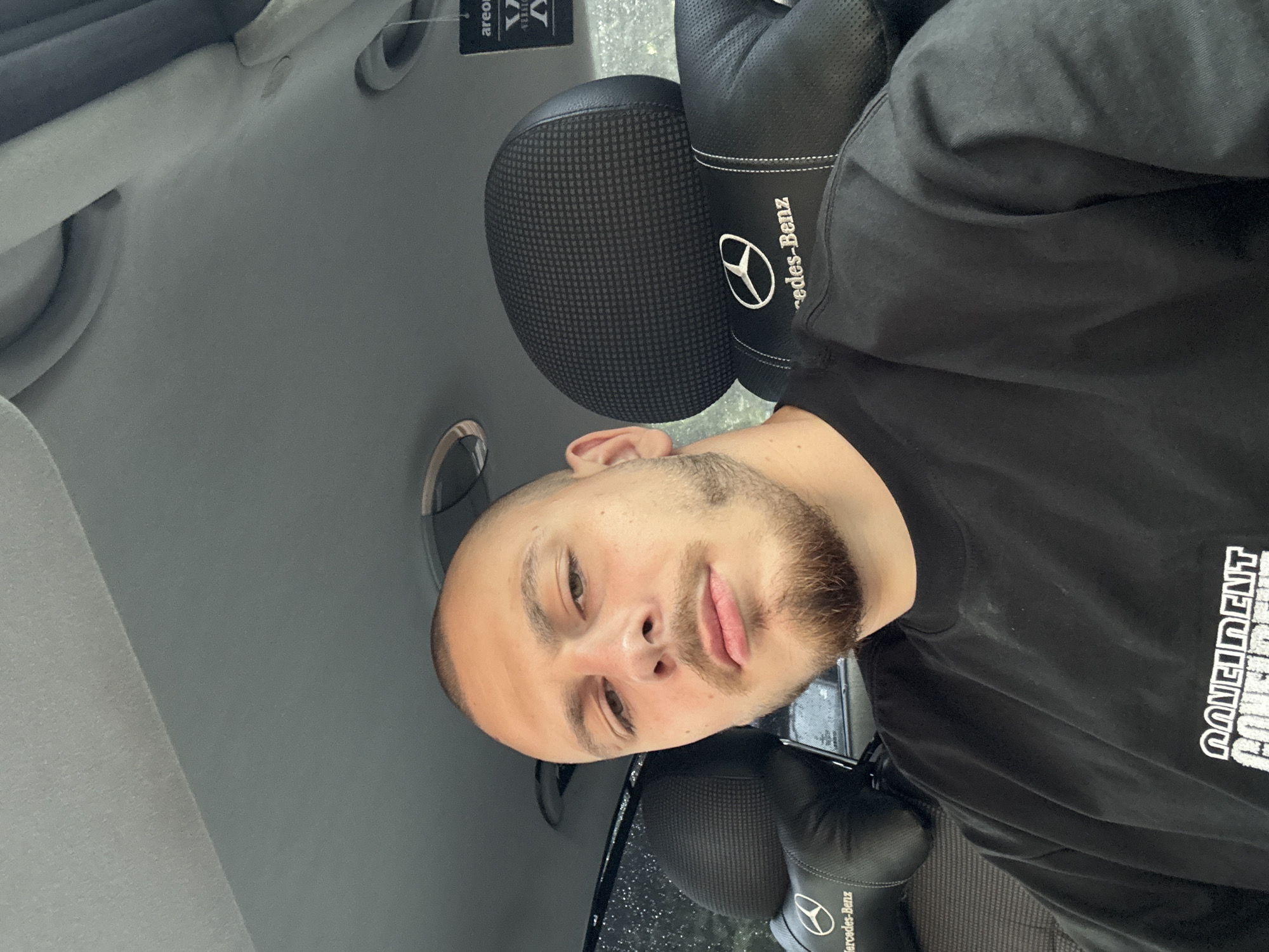 The scene depicts a young adult male inside a vehicle, identified as a Mercedes-Benz based on interior branding, in Stara Zagora, Bulgaria. The man, with light skin, a shaved head, and a light brown full beard and mustache, is wearing a black long-sleeved shirt with partially visible white text reading "CONFIDENT" on the front. He is seated, looking directly at the camera in a selfie-style shot. The interior of the vehicle features black leather seats with perforated accents and patterned fabric headrests, both embroidered with the white Mercedes-Benz logo and text. A black "Areon" brand car air freshener hangs from the overhead area on the passenger side. The vehicle's headliner is light grey. A window on the left side shows blurred exterior elements and possible reflections.