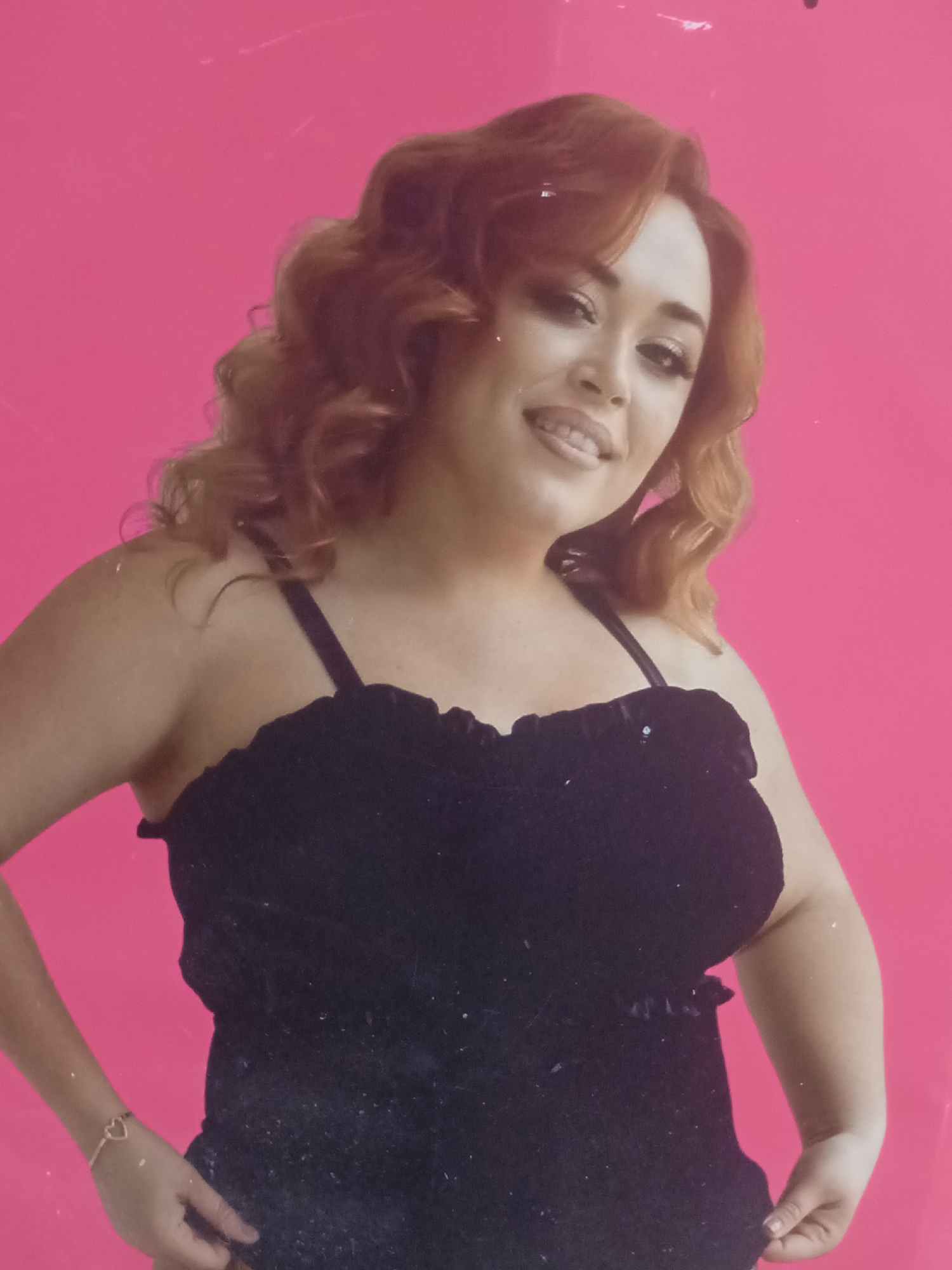 A woman with auburn hair and a black top is posing against a bright pink backdrop. She is smiling and looking towards the camera. A delicate bracelet with a heart-shaped charm adorns her left wrist. The image is lit from the front, casting a soft glow on her face and shoulders. The background is a plain, vibrant pink, with some faint scratches or marks visible. The woman's body is slightly angled, with her left arm bent and her hand near her hip, and her right arm appears to be resting by her side. The overall impression is a posed portrait, likely taken in a studio or similar controlled environment. The image does not contain any elements that would indicate its location in Chernivtsi, Ukraine.
