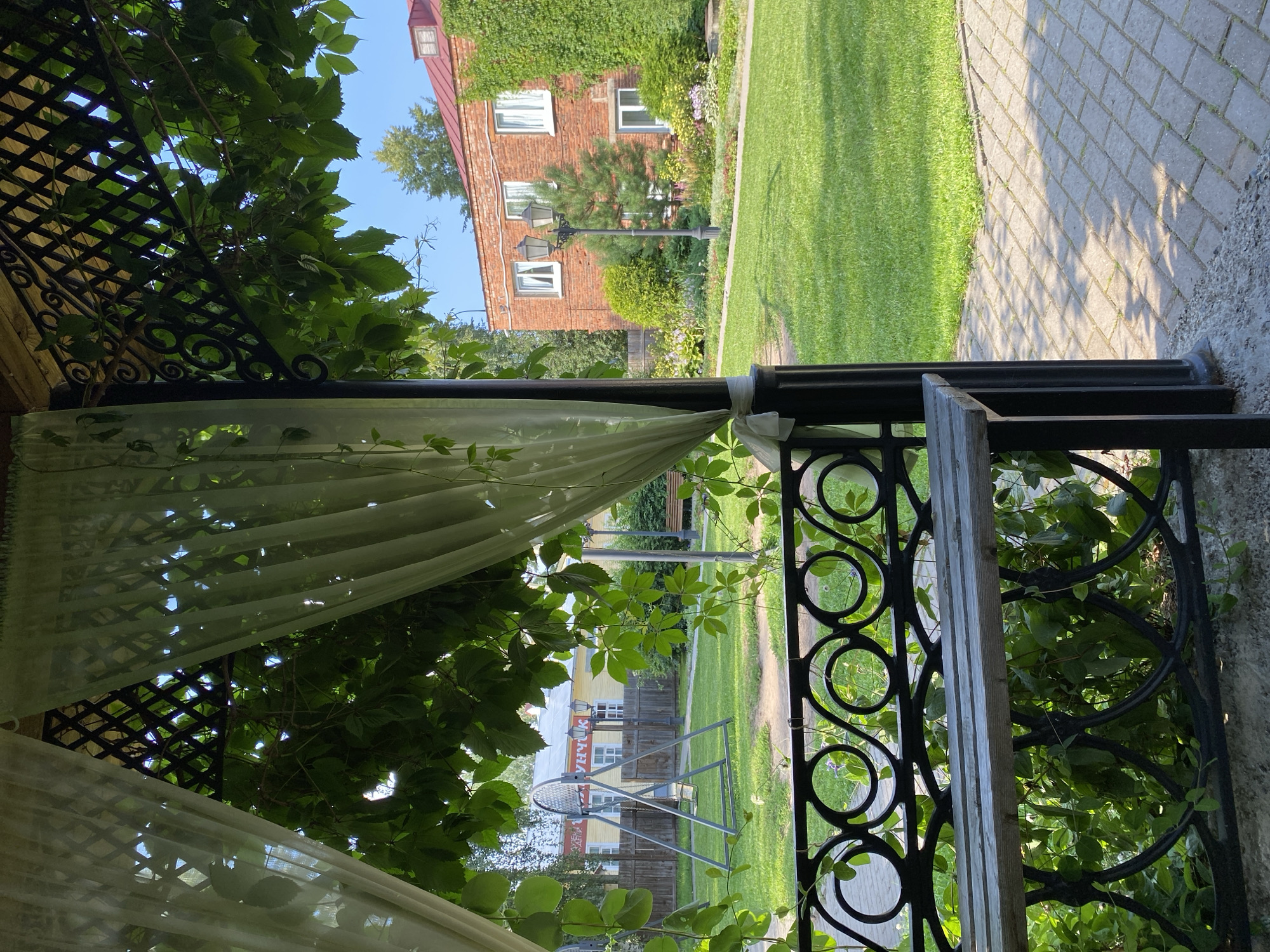 The image depicts a sun-drenched courtyard with a focus on a covered seating area and a manicured lawn. In the foreground, a rustic wooden bench is visible, partially obscured by lush green foliage. The bench is supported by a black metal railing with an ornate, circular design. Sheer white curtains hang from the latticed roof of the covered structure, filtering the sunlight and casting dappled shadows. Beyond the seating area, a vibrant green lawn stretches towards a two-story brick building. The building has a red-tiled roof and white-trimmed windows. A lamppost stands in front of the building, surrounded by a variety of green shrubs and flowering plants. In the middle ground, a swing set is present on the lawn, along with a wooden fence and a yellow building in the distance. The sky is clear and blue. The scene evokes a sense of peaceful, outdoor leisure. This scene is located in Votkinsk, Russia, likely in the vicinity of the Votkinsk State Historical-Architectural Museum-Reserve, which preserves the childhood home of Pyotr Ilyich Tchaikovsky and features similar architectural styles and gardens.