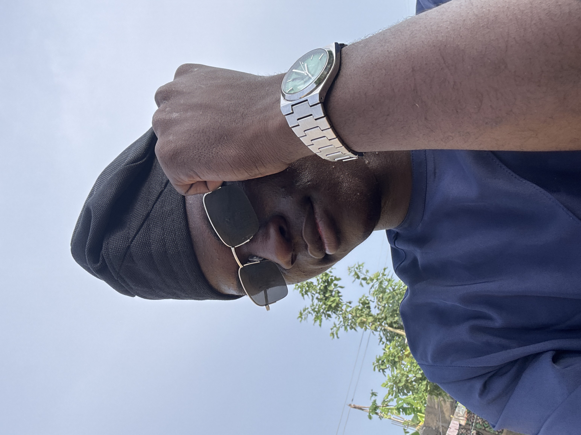 A medium, low-angle shot captures a person with dark skin and a dark headdress wearing sunglasses and a silver watch, likely outdoors in Badagry, Nigeria. The person's right hand is held near their forehead. The background reveals a bright, slightly overcast sky, some green foliage, a utility pole, and a glimpse of a building. The individual is wearing a dark blue top. The scene appears to be captured during daylight hours.