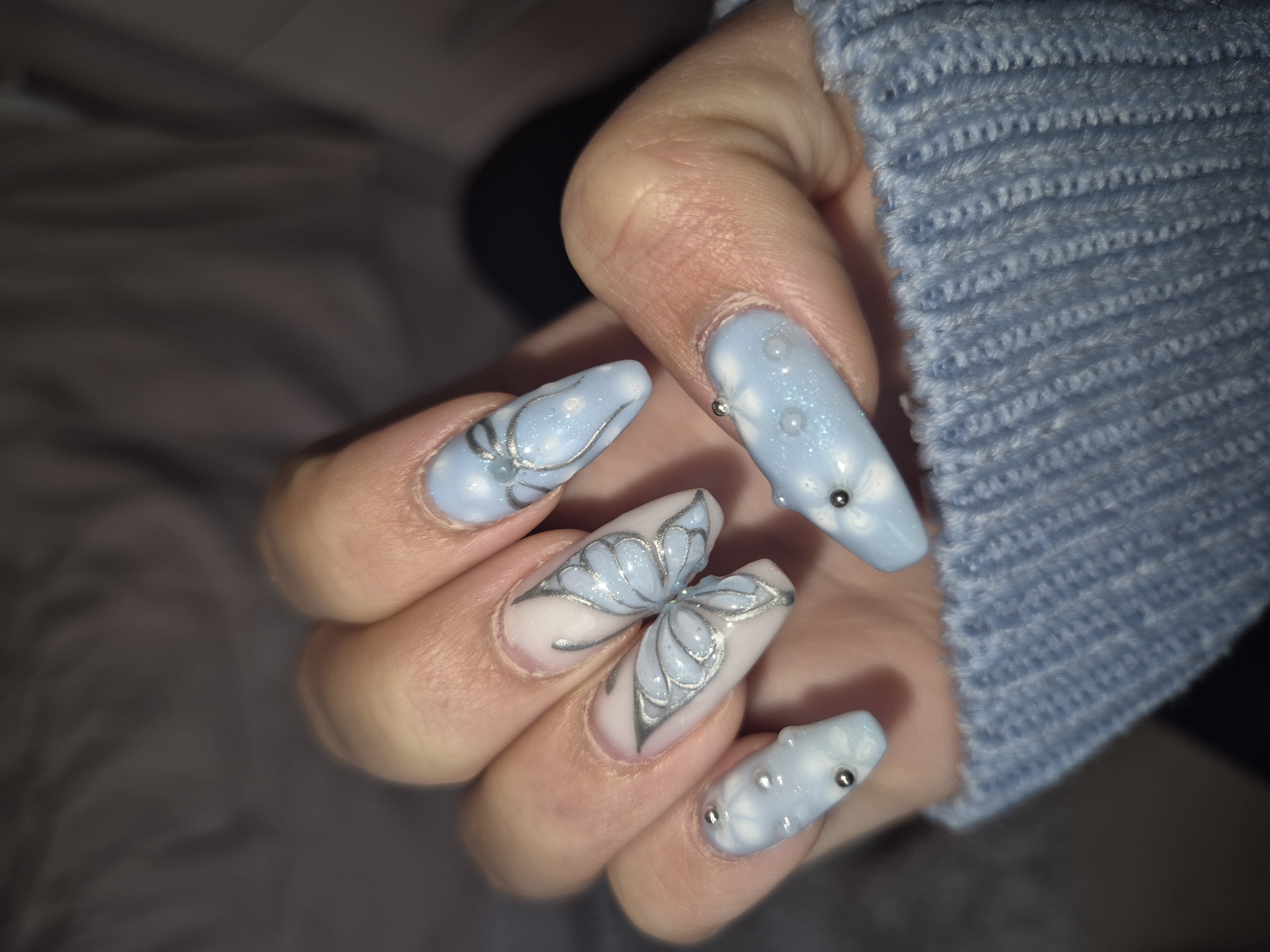 A close-up shows a hand with decorated fingernails against a soft background. The ring finger nail has a white base with a butterfly design outlined in silver. The other nails are painted light blue with silver accents and small white flower-like designs. The person is wearing a blue knitted sweater. The location is not discernible from the image.