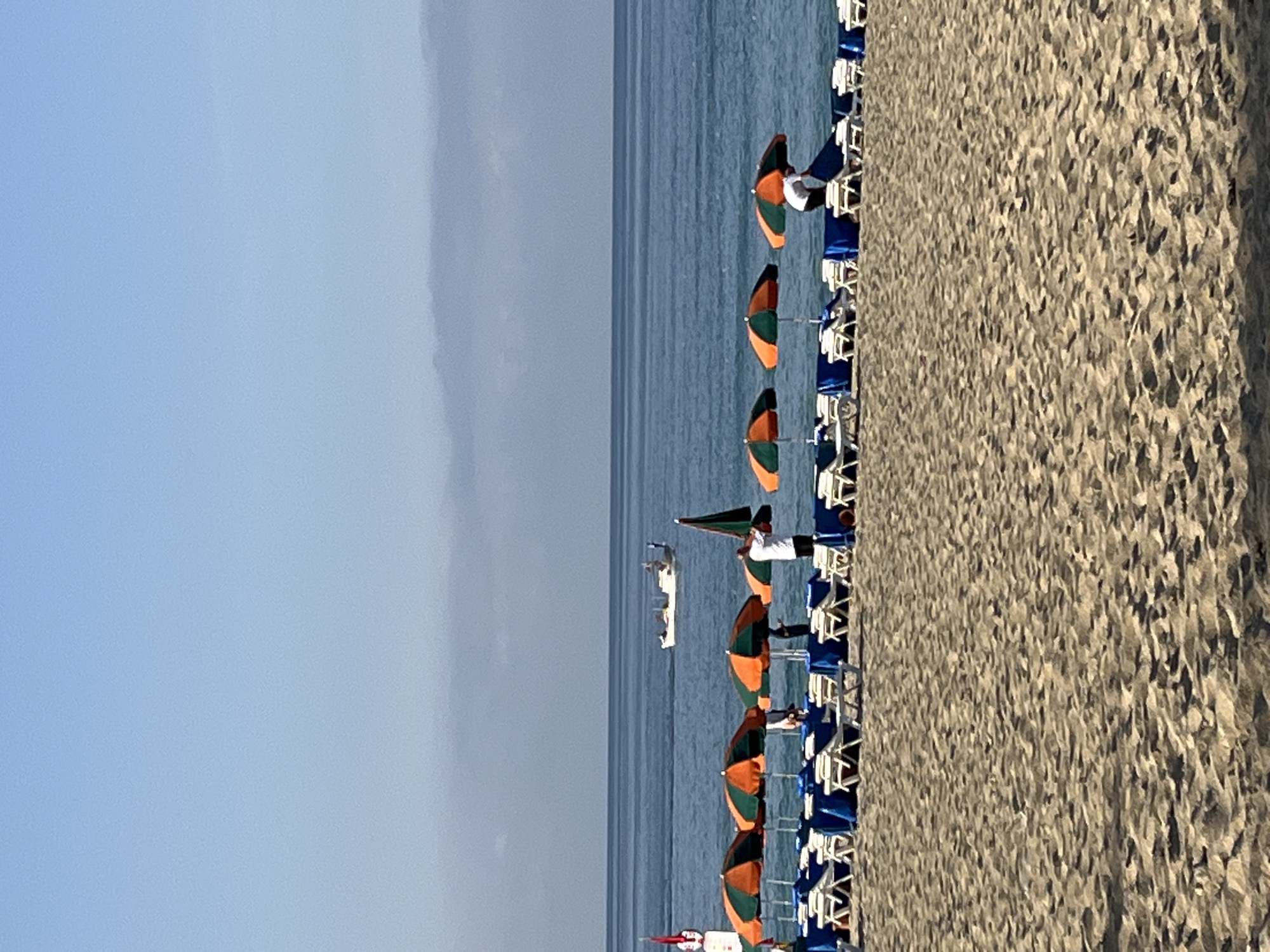 The image shows a beach scene in Alanya, Turkey. The foreground features a sandy beach with rows of lounge chairs and colorful umbrellas. A person in a white shirt is standing near the umbrellas. In the background, there is a blue sea with a boat visible in the distance. The sky is clear and blue.