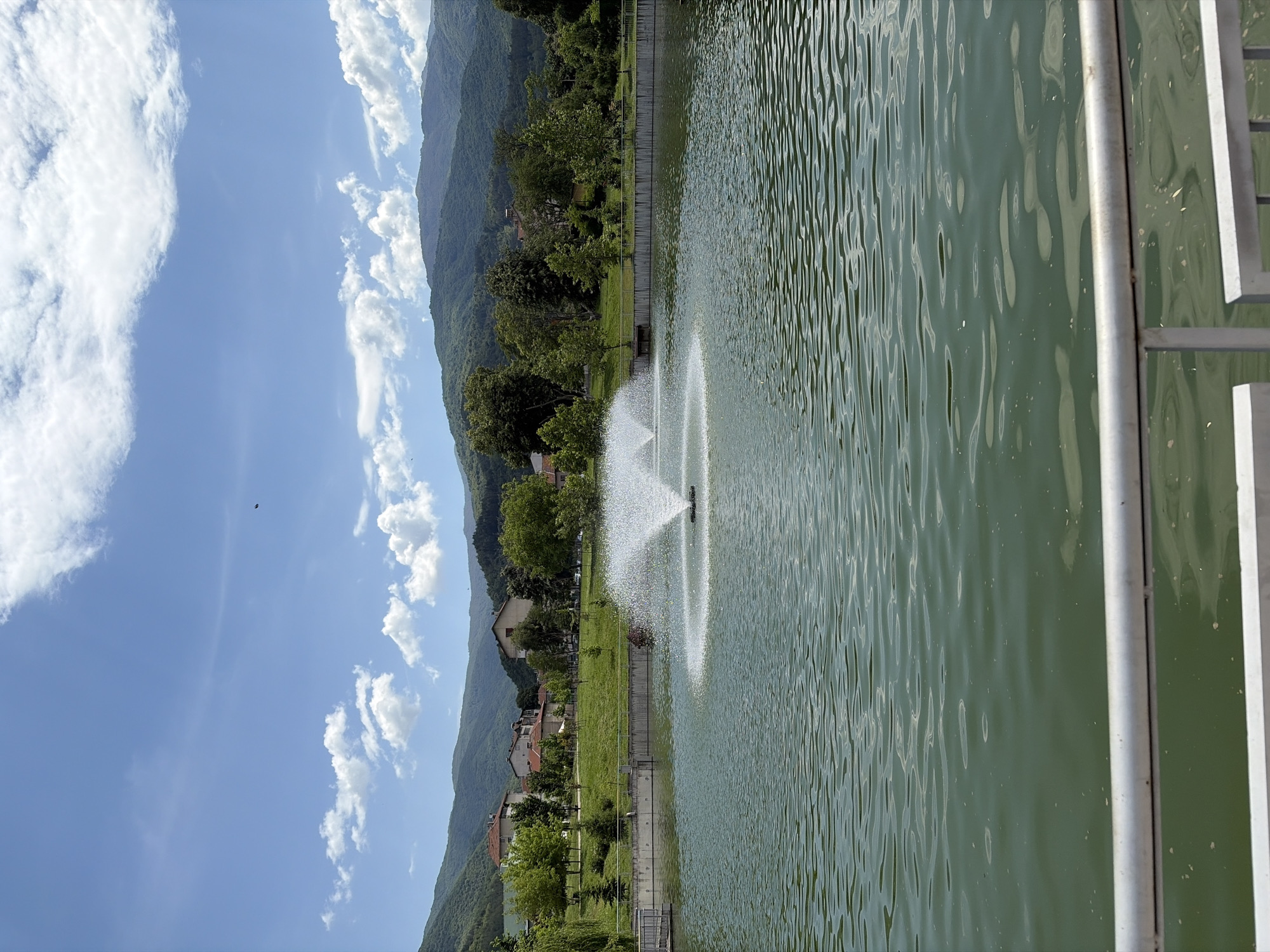 The image shows a body of water, possibly a lake or a large pond, with a fountain in the center. The fountain is spraying water upwards, creating a circular pattern. The water is a greenish color, and there are visible ripples on the surface. Behind the body of water, there is a grassy area with trees and some buildings. Further in the background, there are mountains. The sky is blue with some white clouds. In the foreground, there is a silver metal railing, possibly part of a walkway or bridge next to the water. The image is taken in the city of Unknown, Unknown.