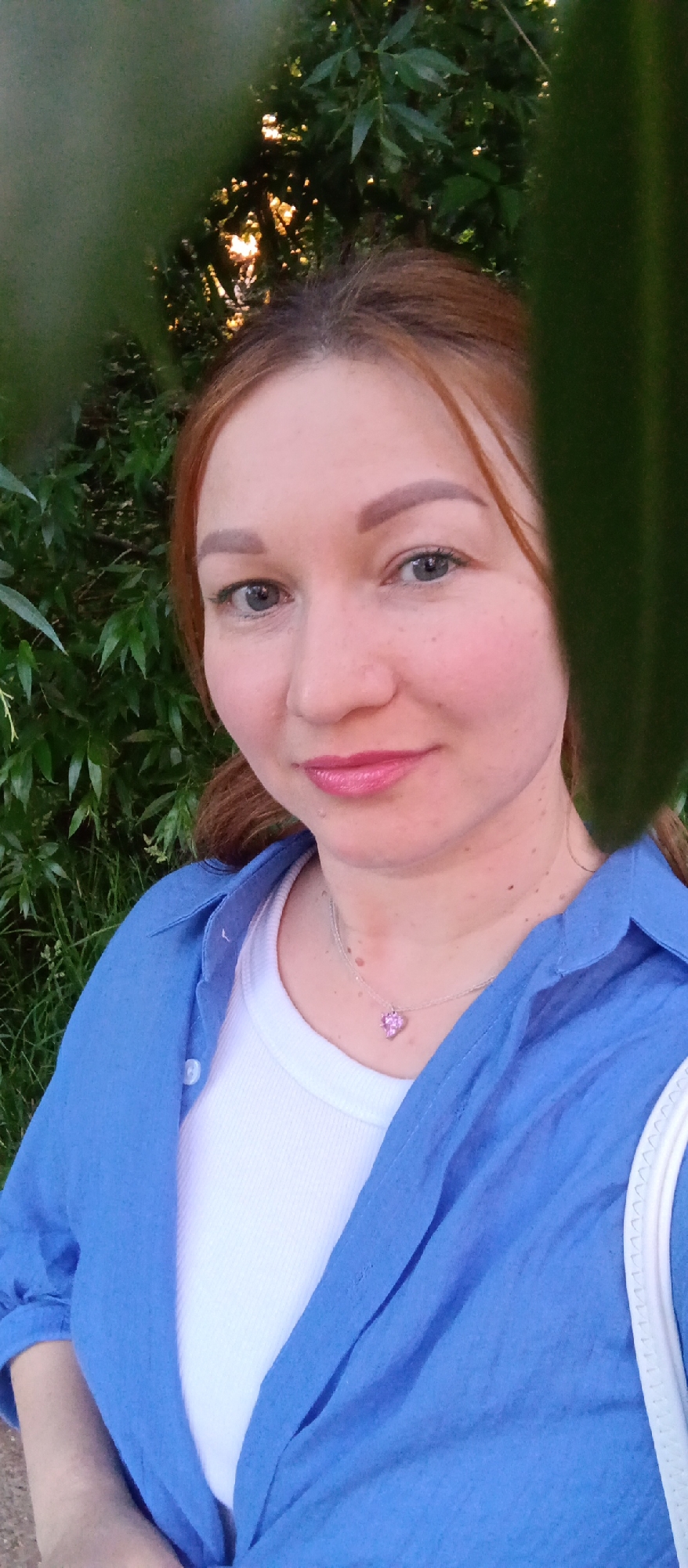 A Caucasian woman, appearing to be in her 30s or 40s with reddish-brown hair and light blue-grey eyes, is captured in a close-up, chest-up outdoor portrait. She is looking directly at the camera with a slight, closed-mouth smile. Her hair, parted centrally, shows darker roots at the crown. Her eyebrows are well-defined and appear light brown. She has subtle freckles visible on her cheeks and nose, and her lips are painted with a glossy pink lipstick. She wears an unbuttoned, long-sleeved blue collared shirt over a white ribbed tank top. Around her neck, a thin silver-colored chain necklace with a small, multifaceted pink heart-shaped pendant is visible. A white strap, possibly from a bag or garment, is partially visible on the far right of the frame. The background consists of dense, dark green foliage, indicative of bushes or trees, with bright, dappled sunlight filtering through the leaves in the upper left and center-right, creating glowing highlights. The foreground is partially obscured by out-of-focus, darker green leaves on both the left and right sides, framing the woman's face. The scene is set outdoors in what appears to be a natural or park-like environment, located in the city of Chaikovsky, Russia.