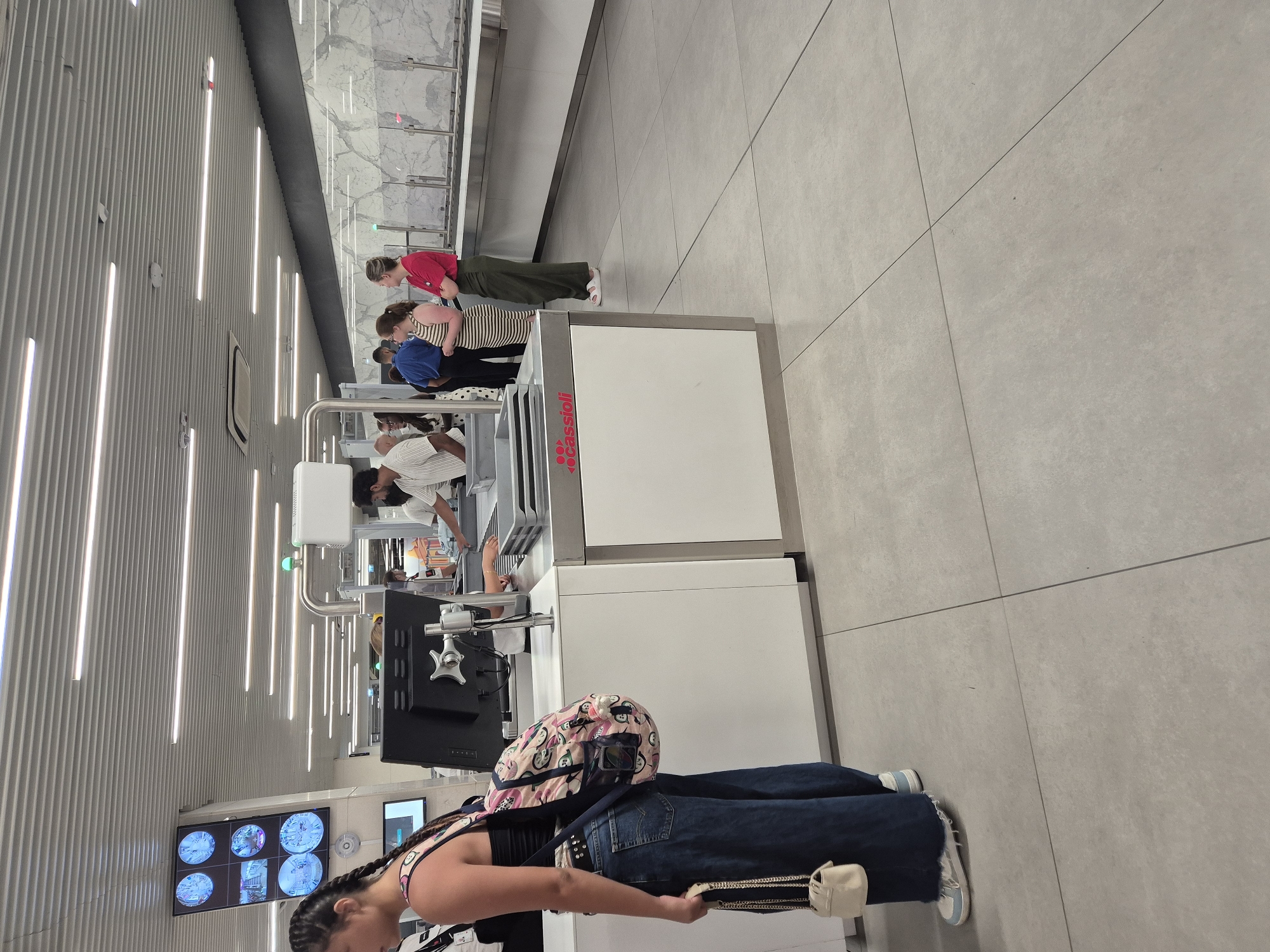 The image depicts an interior scene at Luqa Airport, Malta, likely within the security screening area. In the foreground, a young woman with braided hair is facing away from the camera, wearing a black tank top, dark blue wide-leg jeans, and white sneakers. She is carrying a backpack with a pink and white cartoon pattern and a small white handbag with a chain strap. To her left, a monitor displays multiple circular images, possibly from security cameras.
In the mid-ground, a security checkpoint is visible. A man in a white striped shirt is standing behind a counter, interacting with an individual whose hands are on a conveyor belt. To the right of the man, two women are standing in a line; one is wearing a striped dress, and the other is wearing a red top and green pants. Behind them, a long counter with a marble-patterned backsplash extends into the background. The floor is tiled with light gray square tiles. Overhead, white linear lights illuminate the space, and a metallic ceiling with horizontal slats is visible. The overall environment appears clean and functional, consistent with an airport's security processing area.