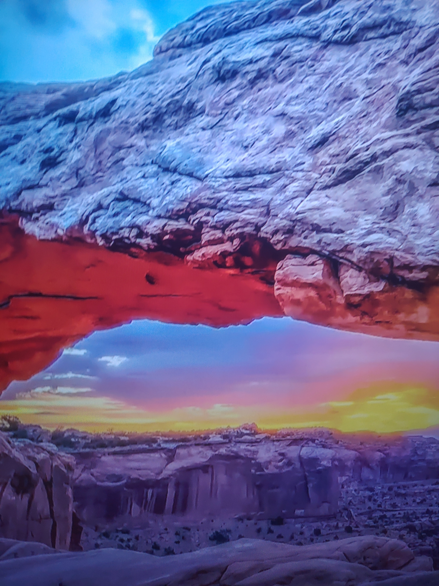 The image captures a sunset view through a natural rock arch. The arch, composed of textured rock, is the dominant foreground element. The top part of the arch is a light gray/blue color, while the part underneath is a more warm red color. In the background, the sky is a gradient of colors from yellow and orange at the horizon to pink and purple above. Below the horizon, there is another rock formation similar in color to the underside of the arch. The location is not specified.