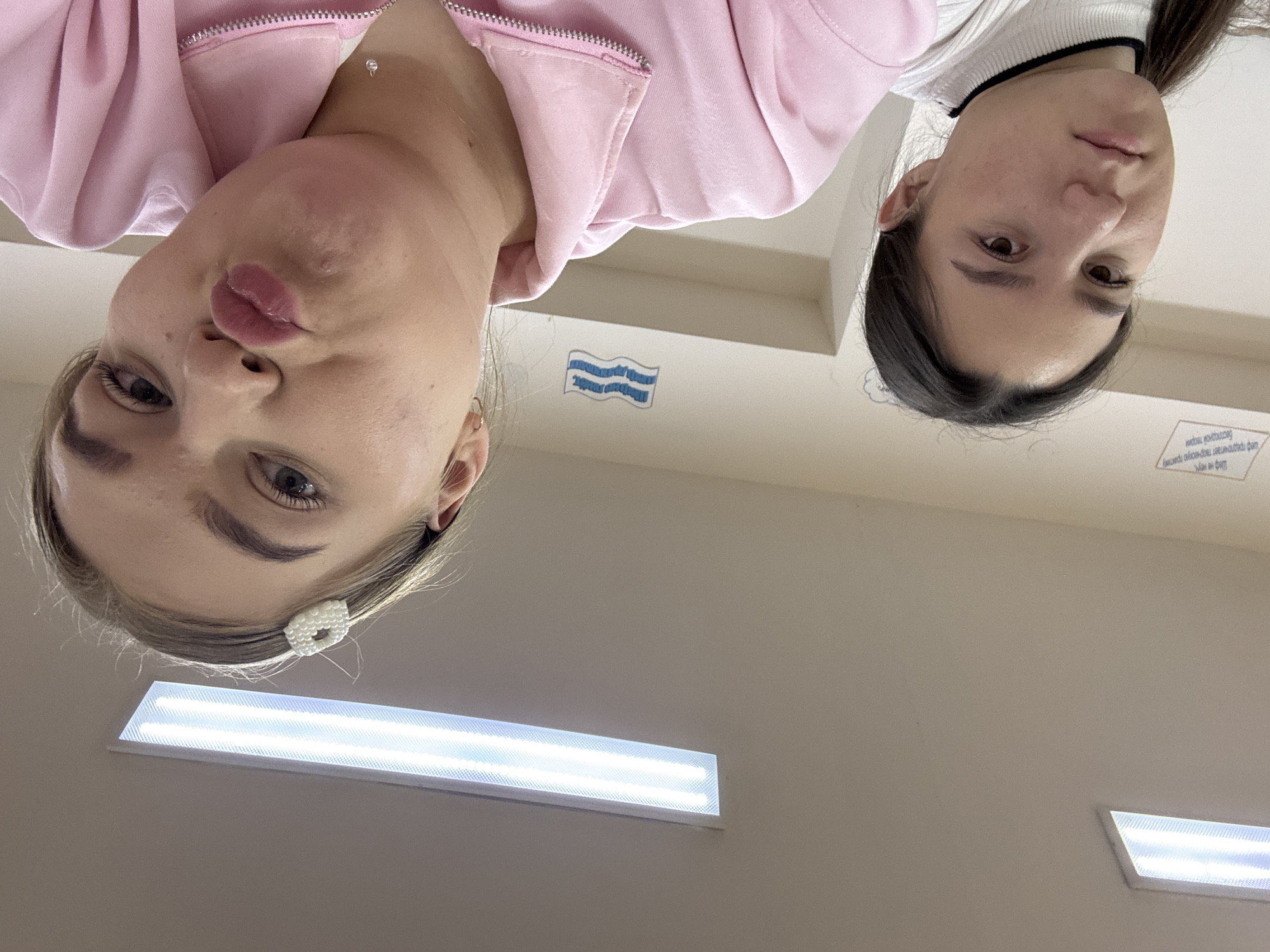 Two young women are looking down at the camera. The woman on the left has blonde hair and is wearing a pink sweatshirt. The woman on the right has dark hair and is wearing a white turtleneck with a black trim. The background appears to be a light beige wall with fluorescent lighting fixtures. There is a small sticker of blue waves on the wall and a small white sign with text in Russian. Based on the presence of Russian text, the location could be in the city of Chaikovsky, Russia.