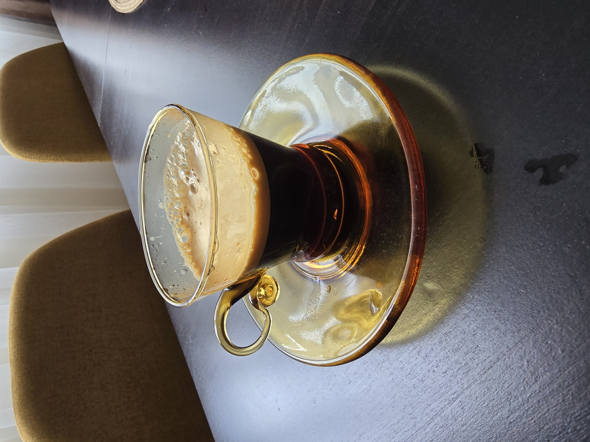 The image shows a glass cup of coffee sitting on a golden-brown glass saucer, placed on a dark, textured surface, likely a table. The coffee has a light, frothy layer on top, with a darker layer beneath. The cup has a small, rounded handle made of the same golden-brown glass. Two mustard-yellow chairs are partially visible in the background, suggesting the location is a cafe or dining area. Two small spots are visible on the table. The scene appears to be indoors, with natural light coming in from the left. The city and location are unknown.