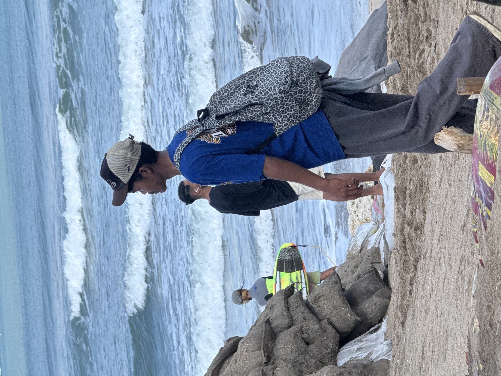 The image depicts a beach scene in Kuta, Indonesia, with the ocean and waves in the background. In the foreground, a young man wearing a blue t-shirt, dark pants, and a baseball cap is walking towards the right. He is carrying a leopard-print backpack. To his left, a person in a black shirt and white shorts is standing, and further left, another person is holding a surfboard with a bright green and yellow design. A large pile of sandbags and rocks is visible on the left side of the image, with a piece of driftwood in the foreground. The sand is a light brown color, and the water is a deep blue.