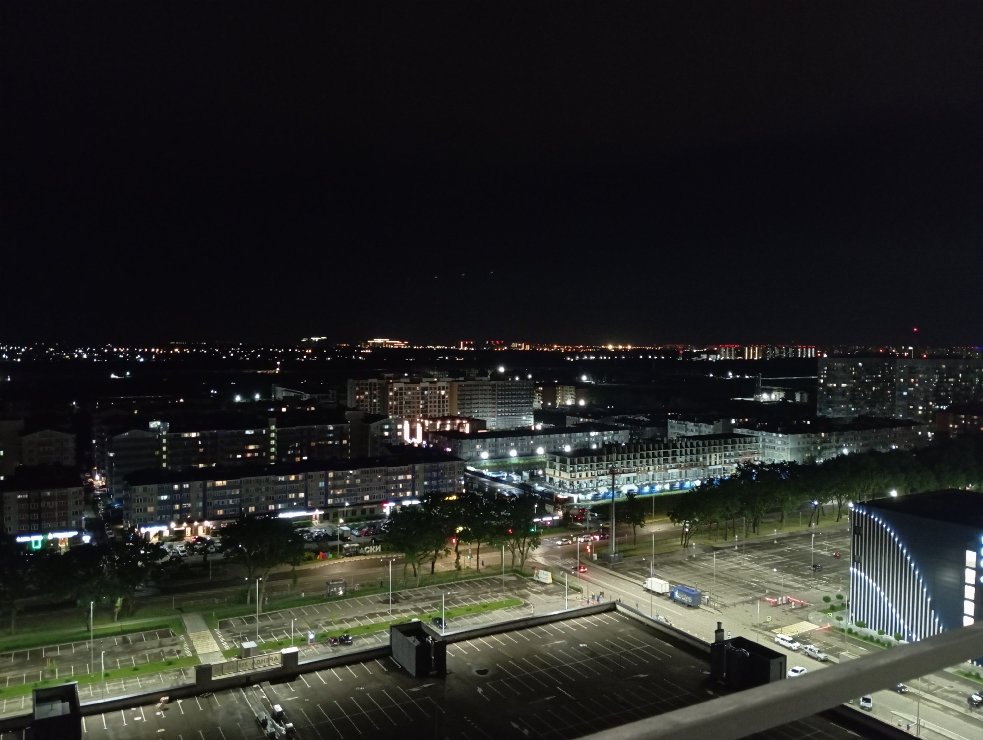 This is an aerial night photograph of a cityscape. The foreground shows an empty parking lot with white lines marking parking spaces. A dark, rectangular structure is situated on the left side of the parking lot. Beyond this parking lot is a road with some vehicles, including a blue truck, and a modern building with vertical white lights on its facade.
Further into the midground, there are several multi-story residential buildings, many of which are illuminated with lights visible through the windows. Trees line a road between the parking lots and the residential buildings. Some of the residential buildings have signs, one of which appears to be in Cyrillic.
The background reveals a more distant cityscape with a denser concentration of buildings and lights, suggesting a larger urban area. The sky is dark and clear, with a faint glow on the horizon from the city lights. The overall scene depicts a typical urban environment at night.