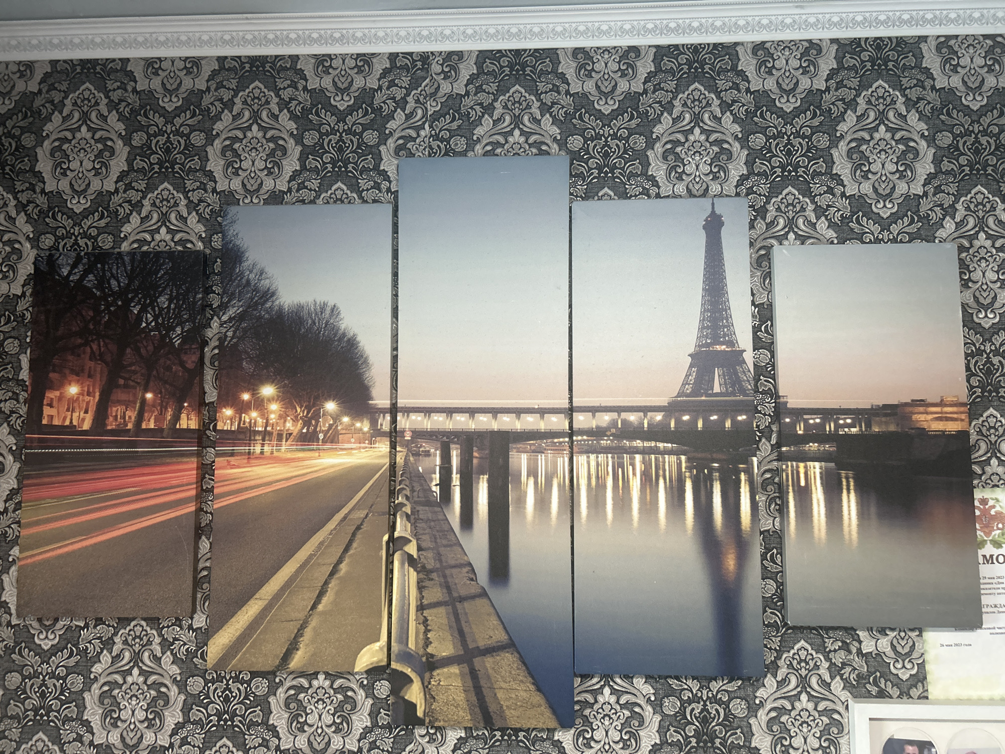 The image displays a multi-panel canvas print of Paris, France, hanging on a wall with damask wallpaper. The canvas print features a scenic view of the Eiffel Tower at dusk or dawn, with a body of water in the foreground reflecting the lights from the city and a bridge. The left panels show a street scene with car light trails and trees. There are no people or objects depicted in the immediate foreground or background of the image, other than the canvas and wallpaper. The photograph is taken from a head-on perspective, with the canvas print occupying the majority of the frame. The question about the city of Kant, Kyrgyzstan is irrelevant to the content of the image.