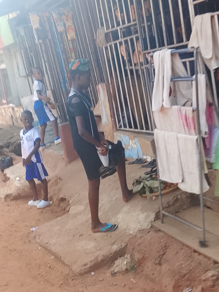 Three children stand outside a house. Two are wearing white shirts and blue shorts with white sneakers. One is standing on a raised cement platform. The other is standing on the ground. The third child, a taller person, is wearing a black dress and a headscarf. They are holding a bag and wearing a single sandal. Behind them is a clothes drying rack with towels, a grilled gate to the house, and a pile of slippers and leaves on the ground.