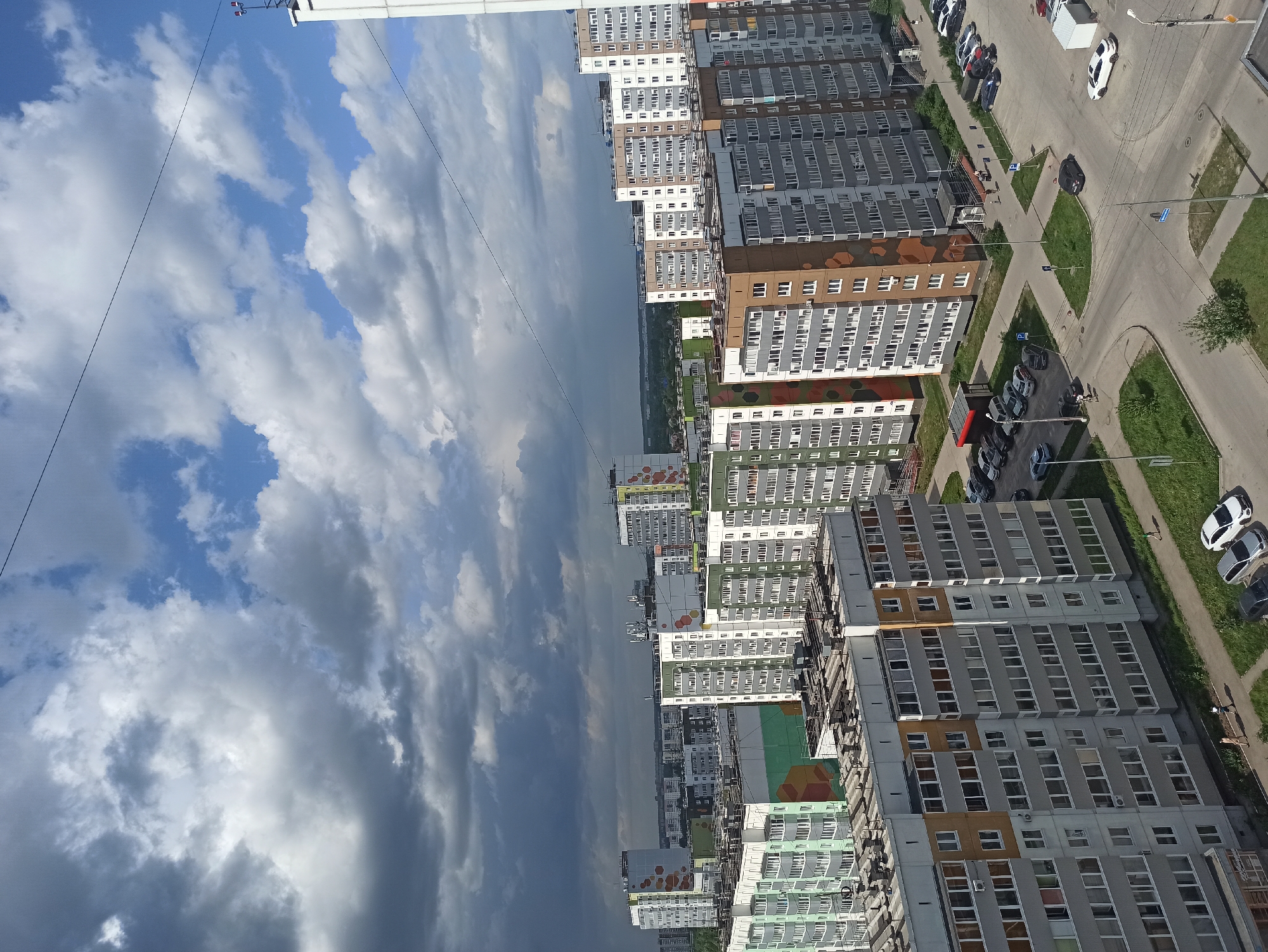 This image shows a cityscape in Unknown, Unknown, from an elevated perspective. A row of multi-story apartment buildings stretches across the frame, interspersed with roads, parking lots, and green spaces. The buildings exhibit a variety of colors and architectural details. Cars are parked in designated areas. Overhead, a partly cloudy sky with white and grey clouds fills a significant portion of the image. A power line or similar cable is visible diagonally across the sky.