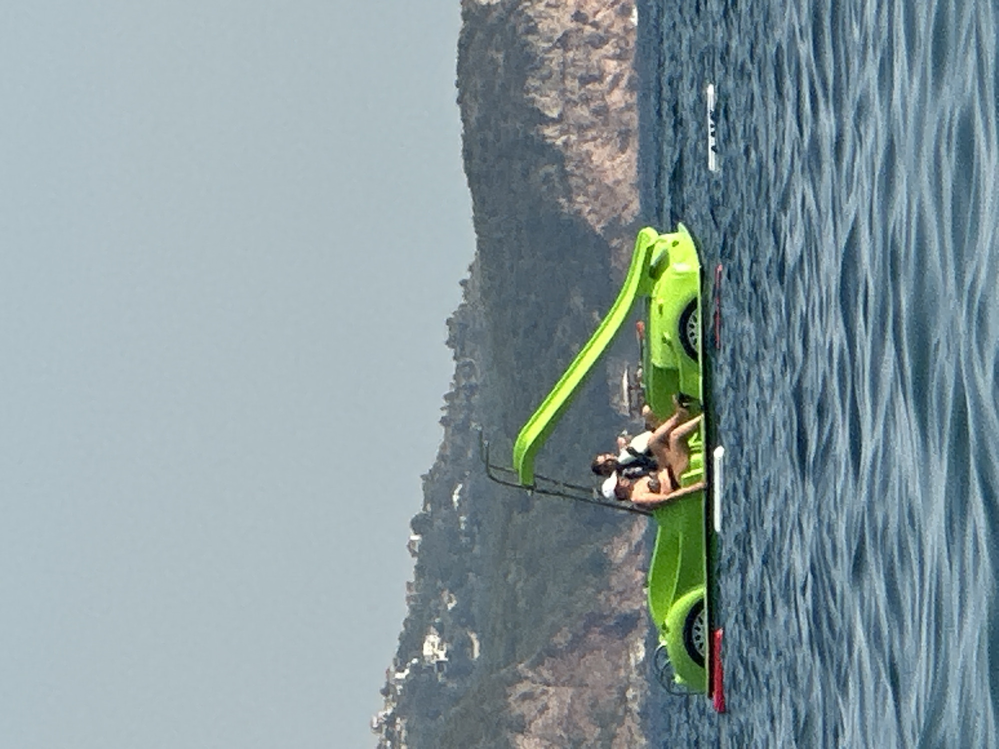 In Boreti, Montenegro, a bright green pedal boat shaped like a sports car is on the water. Two people are seated in the boat. A woman wearing a bikini and a white hat is in the driver's seat. A man wearing a dark shirt is seated next to her. The boat has a slide extending from the back. The water is a deep blue with gentle ripples. In the background, a hilly coastline with buildings and lush green vegetation is visible under a clear, light blue sky.