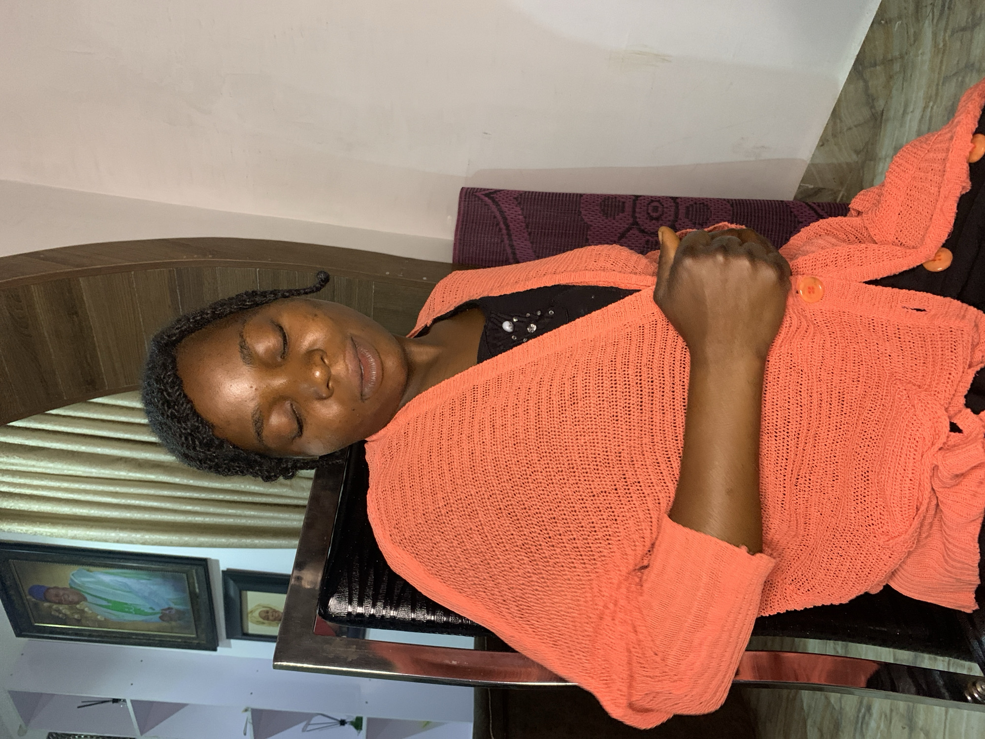 An image taken indoors shows a woman sitting in a chair with her eyes closed. She is wearing a black shirt and an orange knitted cardigan. The cardigan has three buttons down the front. The woman has dark skin and braided hair. The background includes a framed portrait on the wall, gold curtains, and a patterned fabric on the chair. The lighting suggests it is daytime. The location is identified as the city of Jos, Nigeria.