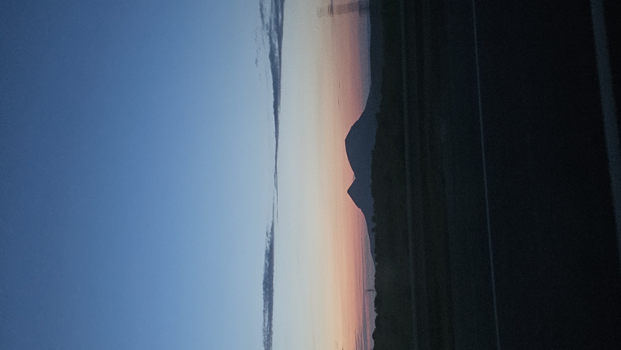 A silhouette of two rounded mountains is visible against a gradient sky transitioning from deep blue at the top to pale orange and pink near the horizon. A dark, silhouetted foreground suggests a landscape, possibly including trees or hills, with a paved road or highway in the lower portion of the image, marked by a white line. The scene appears to be captured at dusk or dawn.