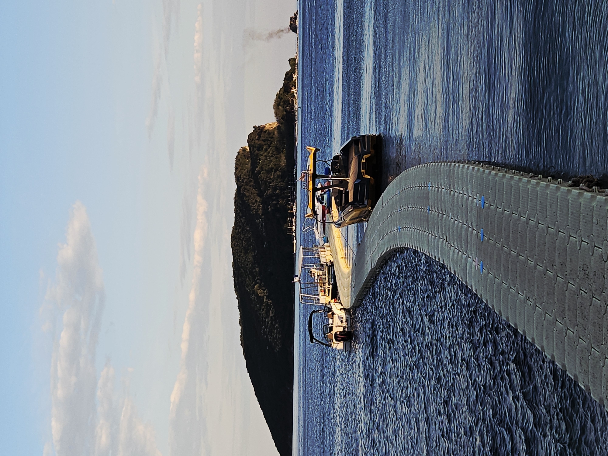 Here is a description of the image.
The image shows a tranquil scene with a calm body of water extending to the horizon. Several boats are docked along a floating pier that curves into the water. The sky is a mix of light blue with soft, scattered clouds. Lush greenery covers a hill or mountain in the background. It is difficult to provide specific location details without additional information.