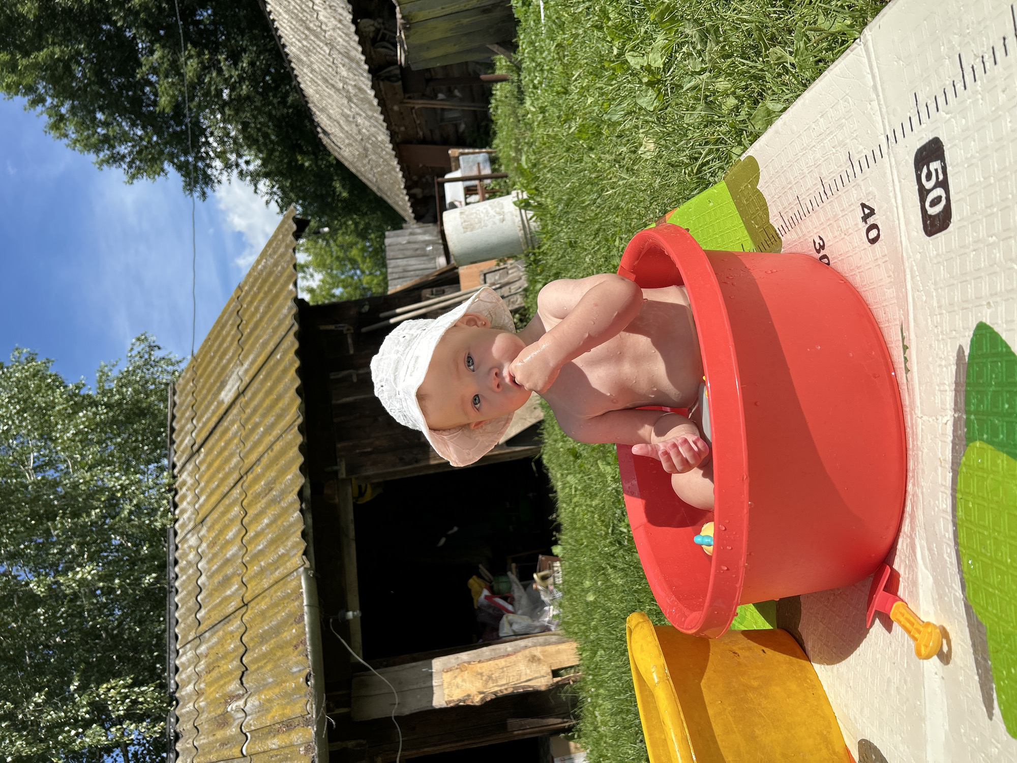 A baby wearing a white sunhat sits in a bright red plastic basin outdoors on a sunny day. The baby is looking at the camera with a curious expression. The basin appears to be part of a play area, as a yellow slide is visible next to it. A mat with measurement markings, ranging from 30 to 50, is placed on the grass next to the basin. The background shows a rustic setting with a weathered wooden structure, possibly a shed or outbuilding, with a corrugated metal roof. Lush green grass and trees frame the scene, and the sky is blue with some scattered clouds. The overall impression is one of a warm, summer day spent playing outdoors in a rural environment.