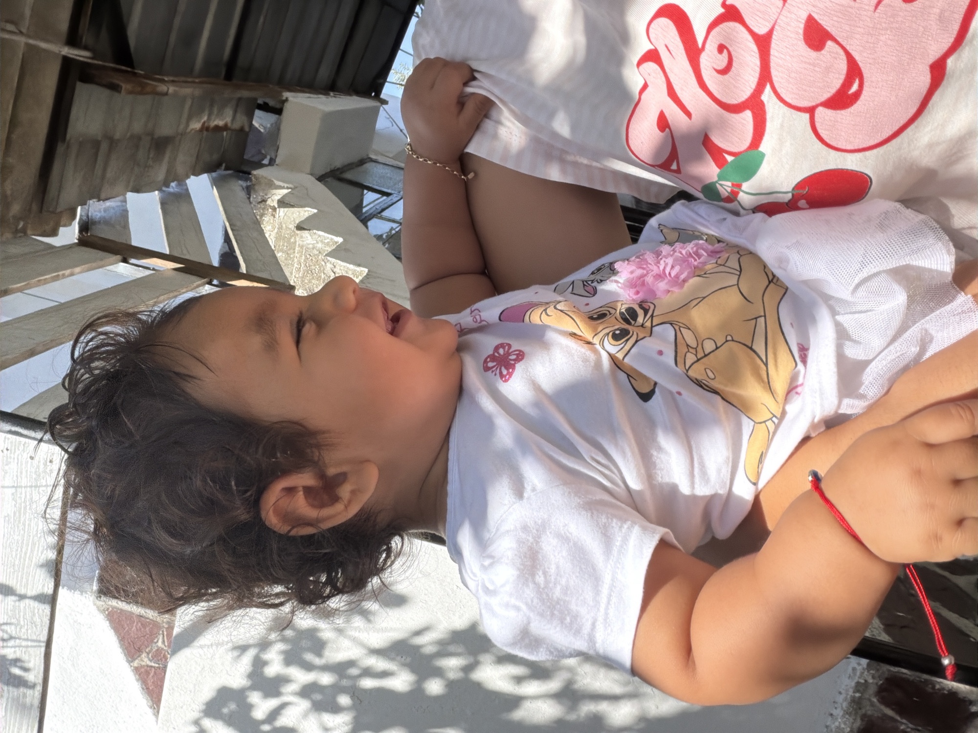 In Razhevo Konare, Bulgaria, a young child with dark, curly hair is seated outdoors, bathed in sunlight. The child is wearing a white t-shirt adorned with a cartoon character and a pink floral embellishment, along with a white skirt. A red string bracelet with a silver bead is visible on the child's left wrist. The child is positioned in front of an adult, whose arm and part of a t-shirt with a cherry design are visible. The background shows a white wall with dappled light and shadows, suggesting the presence of foliage, and a rustic structure with wooden beams and corrugated metal roofing. The overall setting appears to be a residential outdoor area.